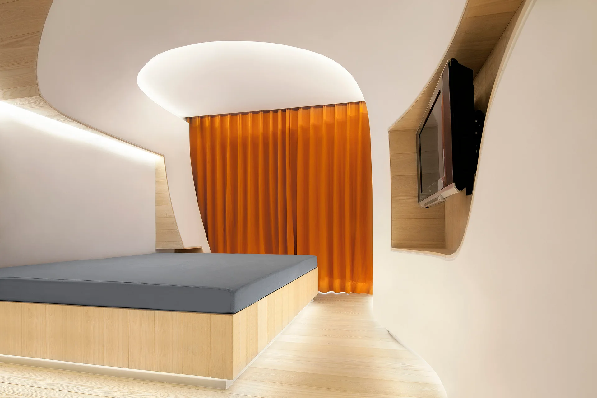 A minimalist bedroom features curved white walls and ceiling, a light wood platform bed, an integrated television niche, and a bright orange curtain.