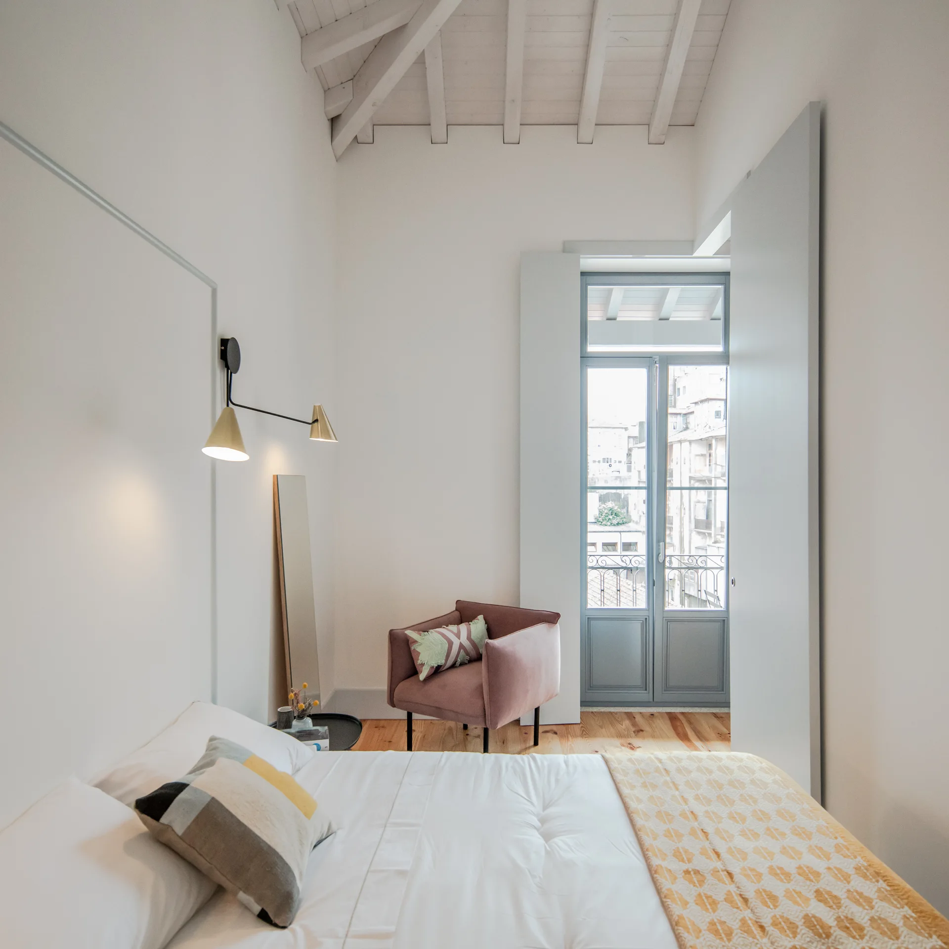 Bright bedroom with a white bed, a dusty pink corner armchair, and a tall mirror under a white-painted vaulted wood beam ceiling.