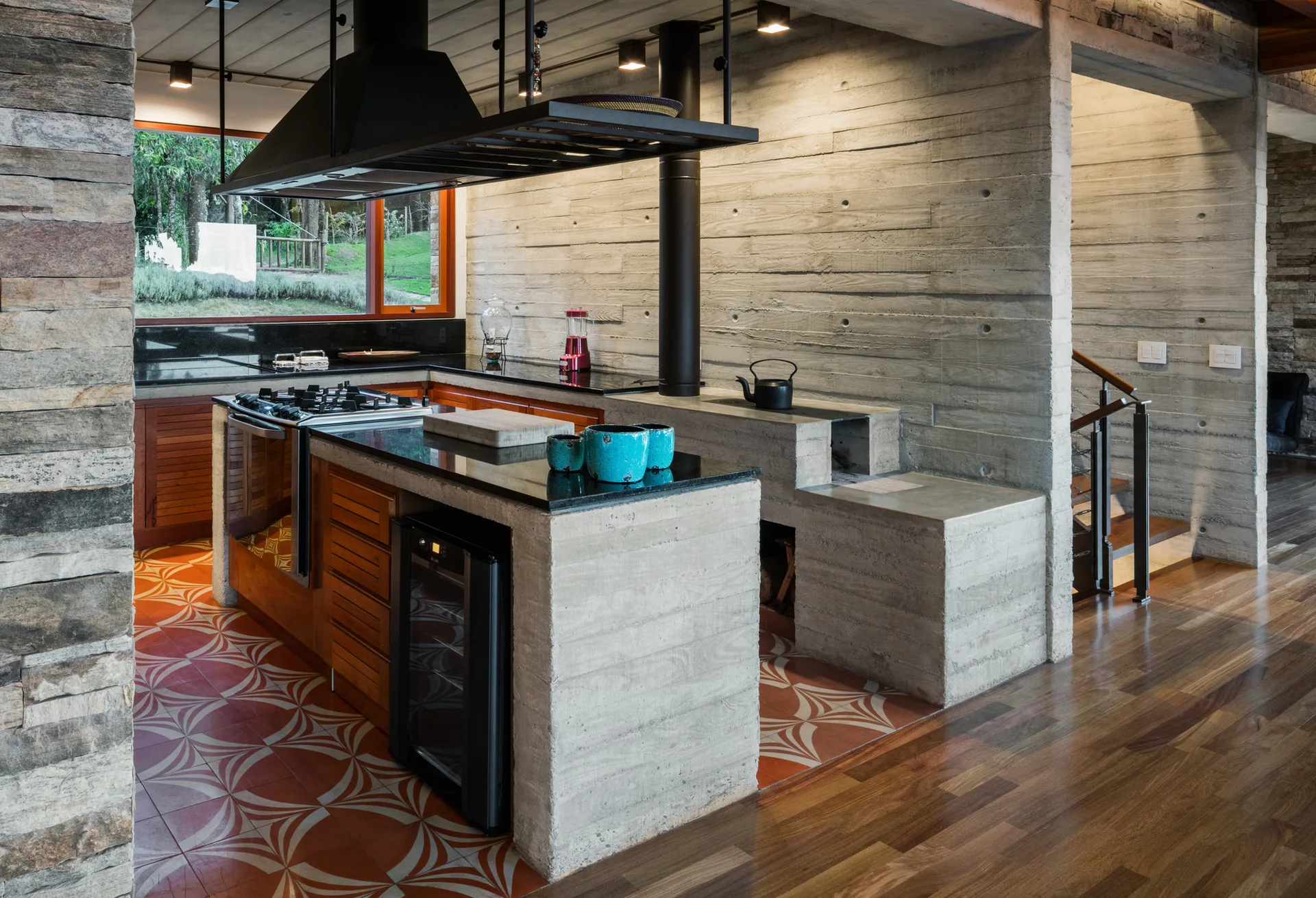 A modern kitchen features a board-formed concrete island with a gas cooktop, black countertops, and geometric patterned floor tiles.
