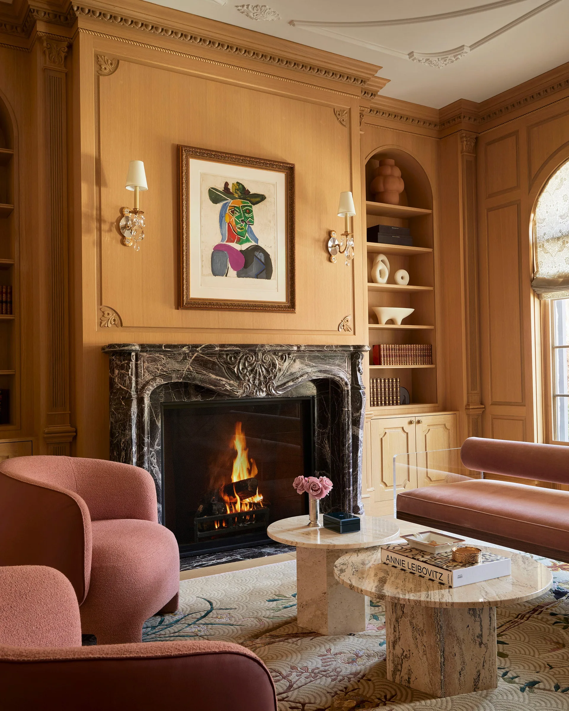 A warm wood-paneled living room featuring an ornate marble fireplace, arched built-in shelving, pink armchairs, and round stone nesting coffee tables.