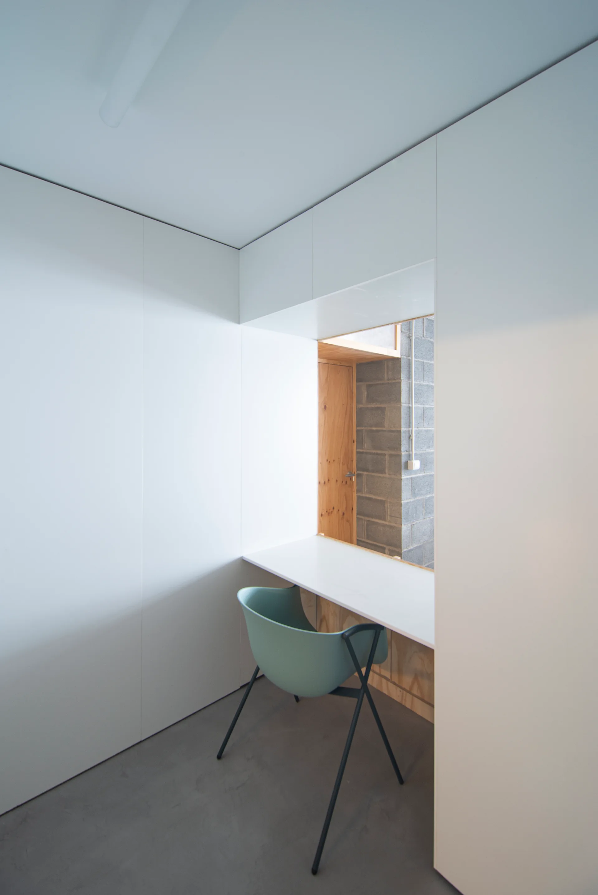 A minimalist white built-in desk nook features a light green chair, smooth gray flooring, and an opening showing a concrete block wall beyond.