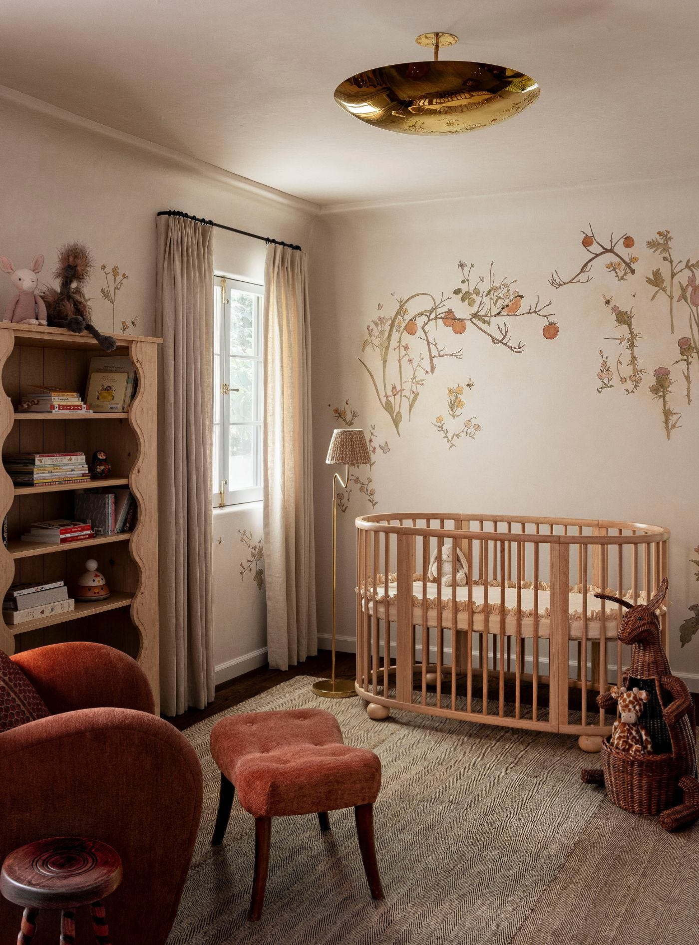 A cozy nursery featuring a light wood oval crib, a scalloped bookshelf, a terracotta armchair, and botanical illustrations on cream walls.