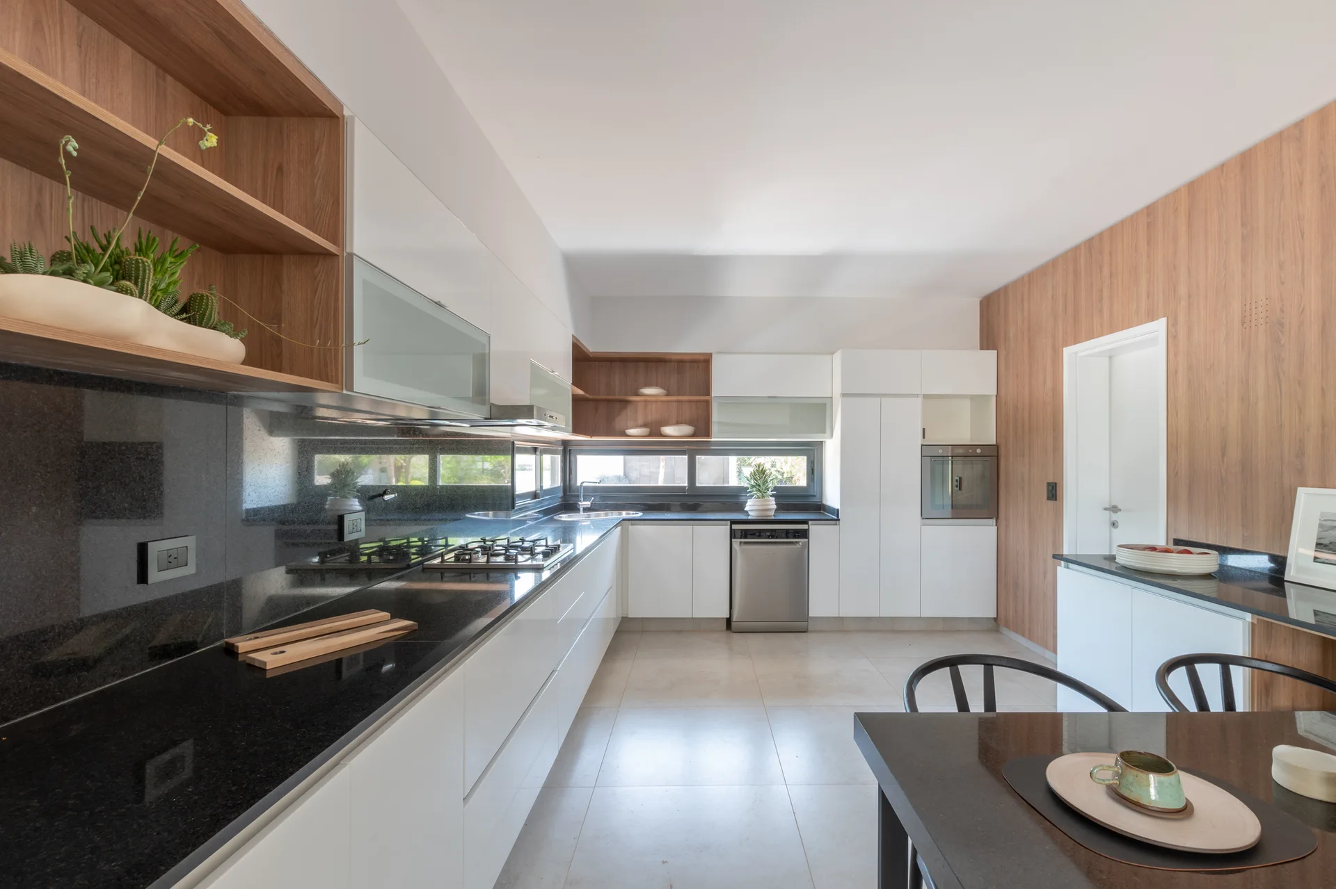 A modern kitchen with white slab cabinetry, black countertops, warm wood-toned open shelves, and a horizontal strip window.