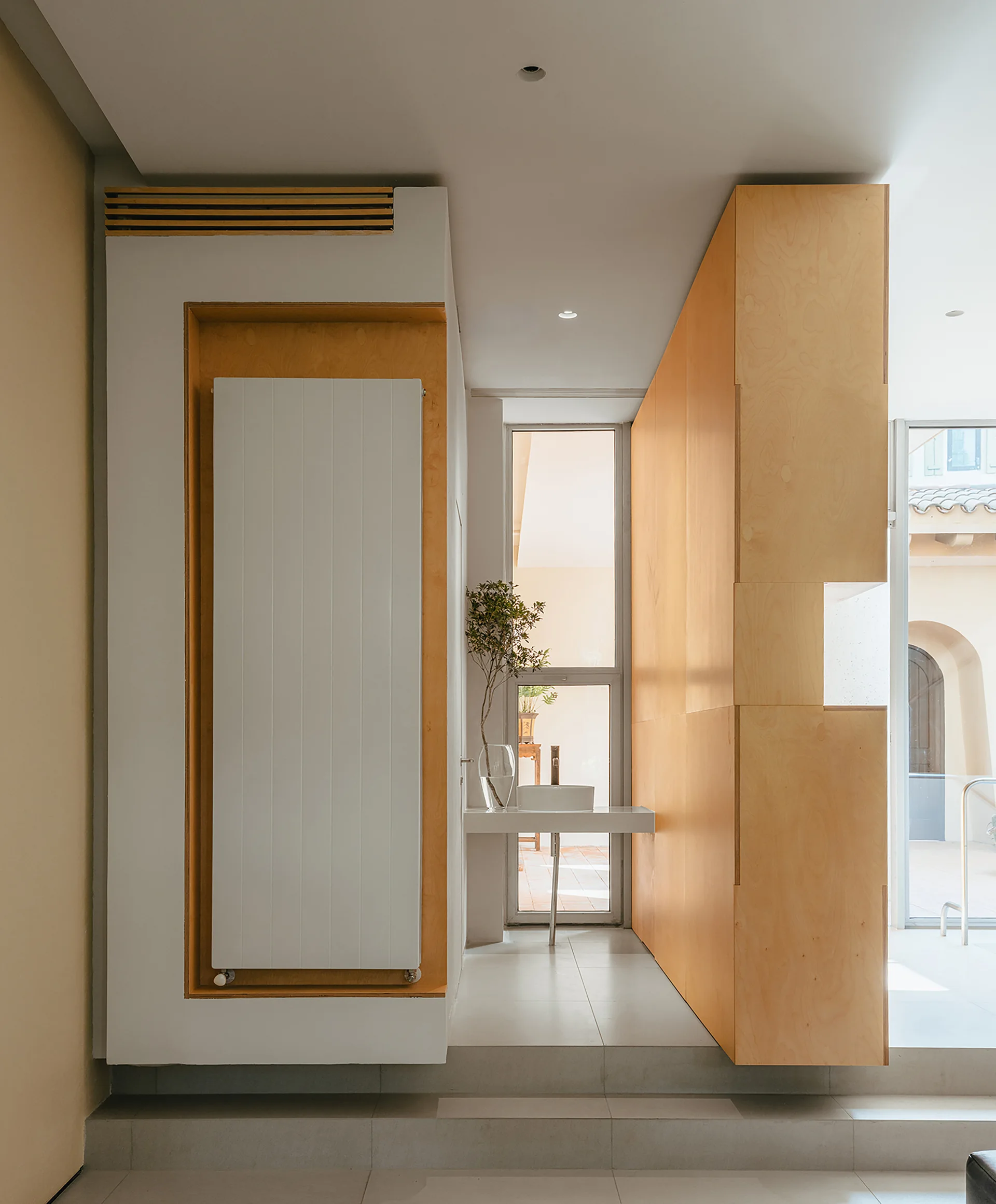 A minimalist bathroom featuring a tall plywood partition wall, a floating sink placed before a window, and a recessed vertical radiator.