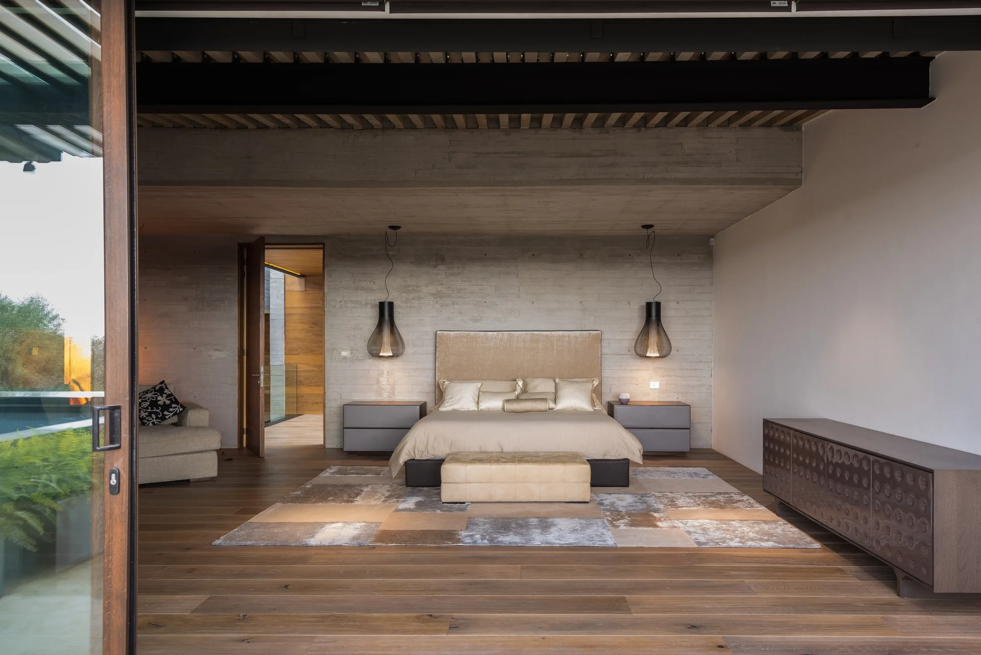 A modern bedroom features a neutral bed set against a gray board-formed concrete wall, flanked by gray nightstands and hanging wire pendant lamps.