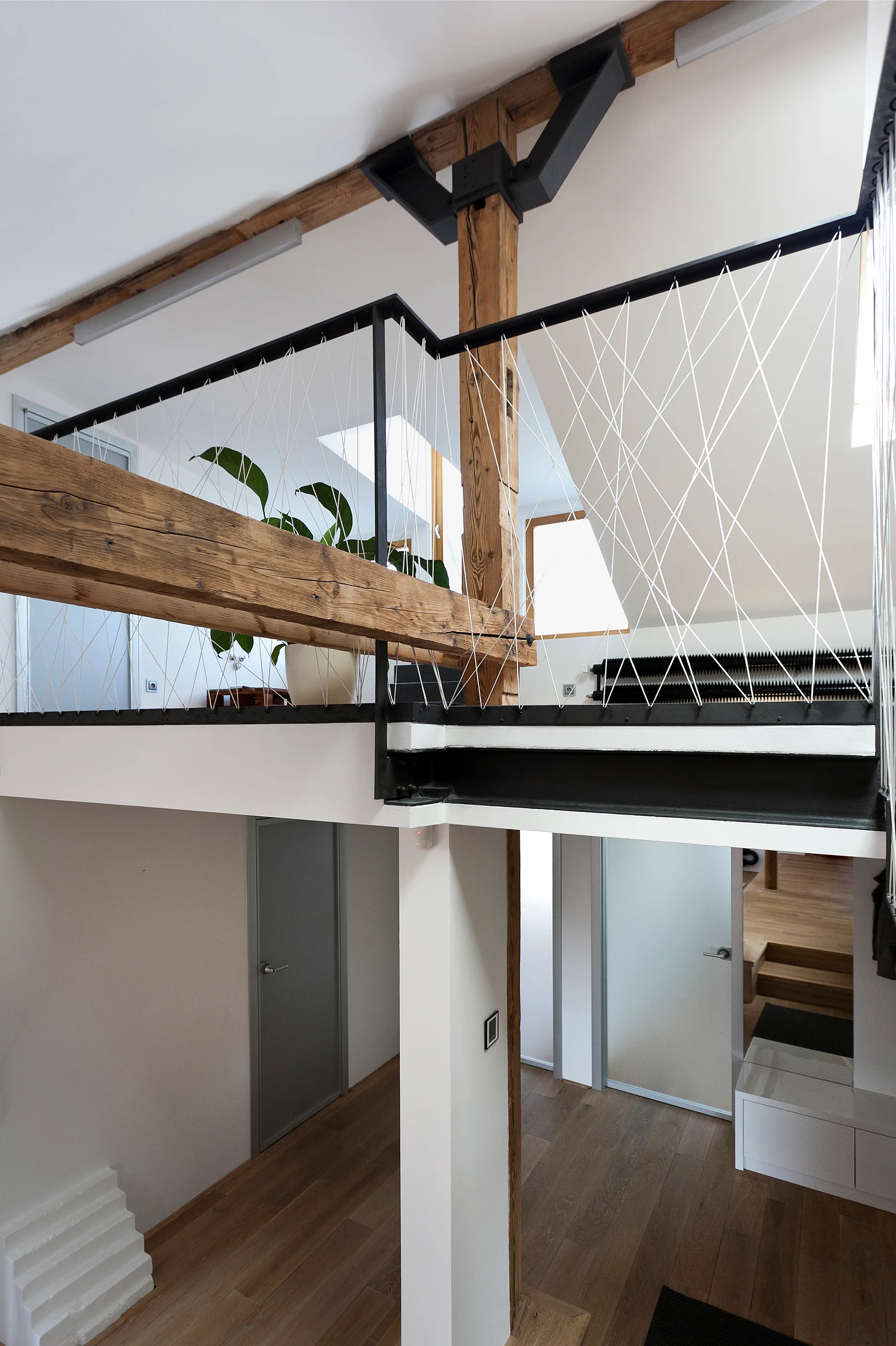 An elevated interior mezzanine features a geometric white cord railing alongside rustic wood columns and beams overlooking a lower level.