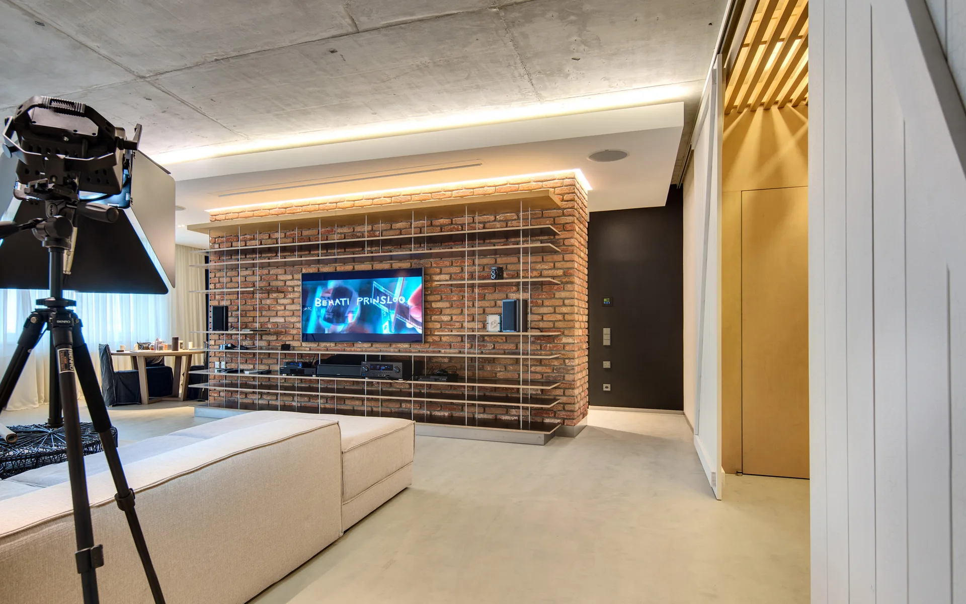 A modern living room features an exposed brick feature wall with mounted shelving, a concrete ceiling with cove lighting, and a beige sofa.