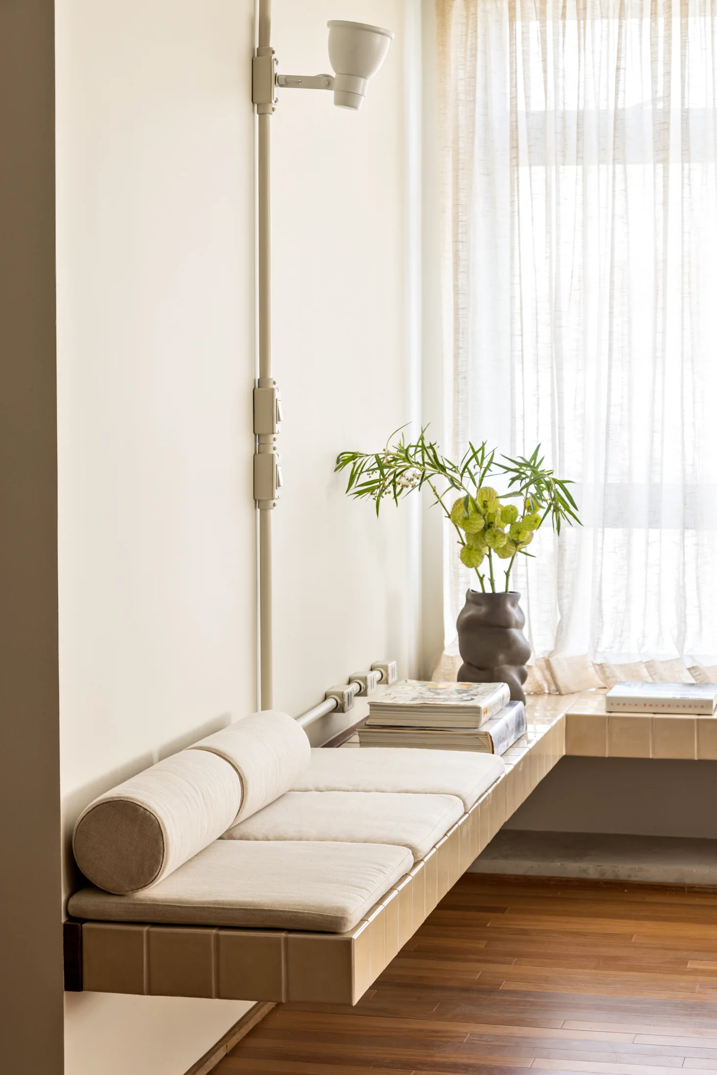 A cream-colored tiled built-in bench is topped with soft cushions and a bolster pillow next to a window with sheer curtains.