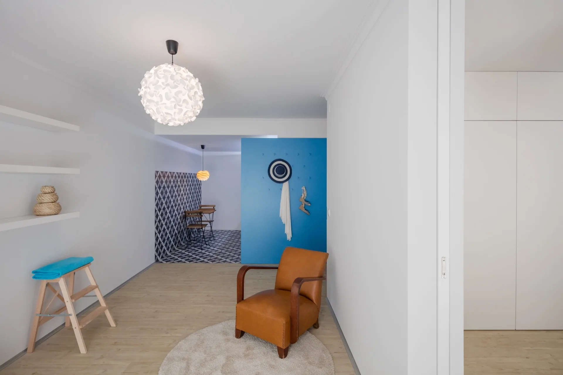 A tan leather armchair sits on a round rug in a white room featuring a bright blue accent wall and a dining nook with geometric floor tiles.