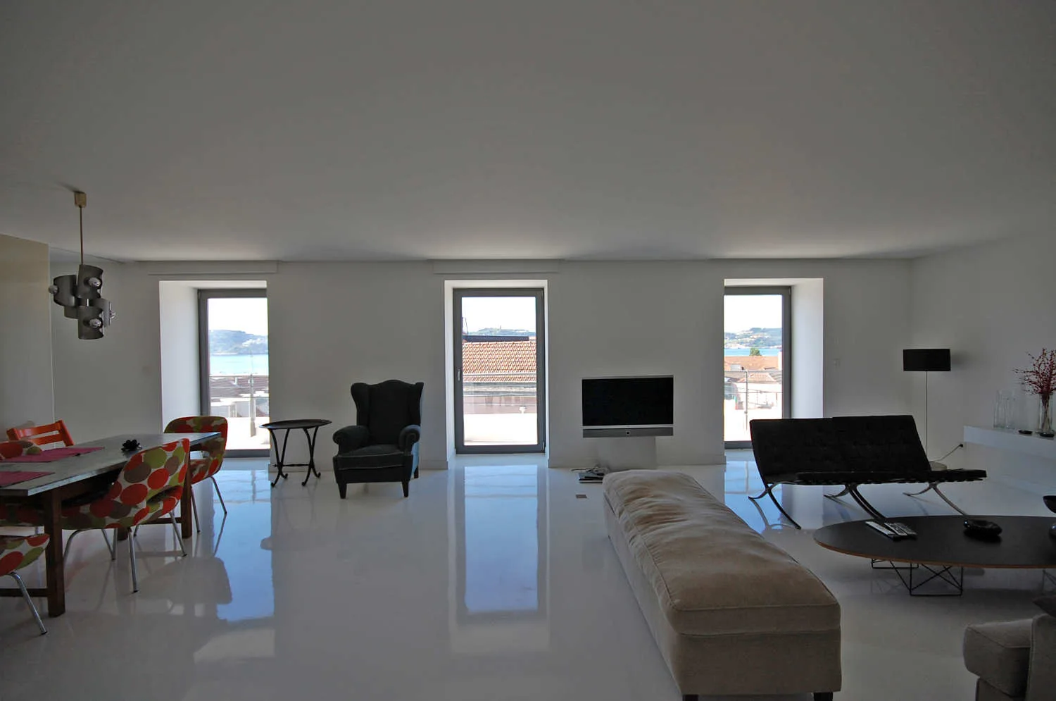 An open-plan living room features high-gloss white flooring, three vertical windows, and a mix of modern black and colorful patterned seating.