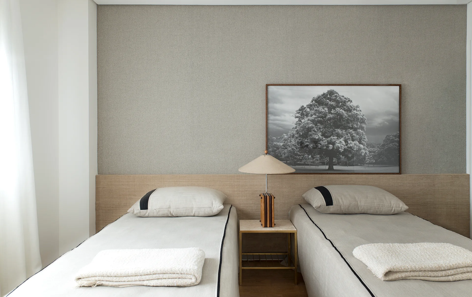 Two twin beds with beige linens are framed by a long woven headboard, with a small nightstand and a framed tree photograph on the wall.