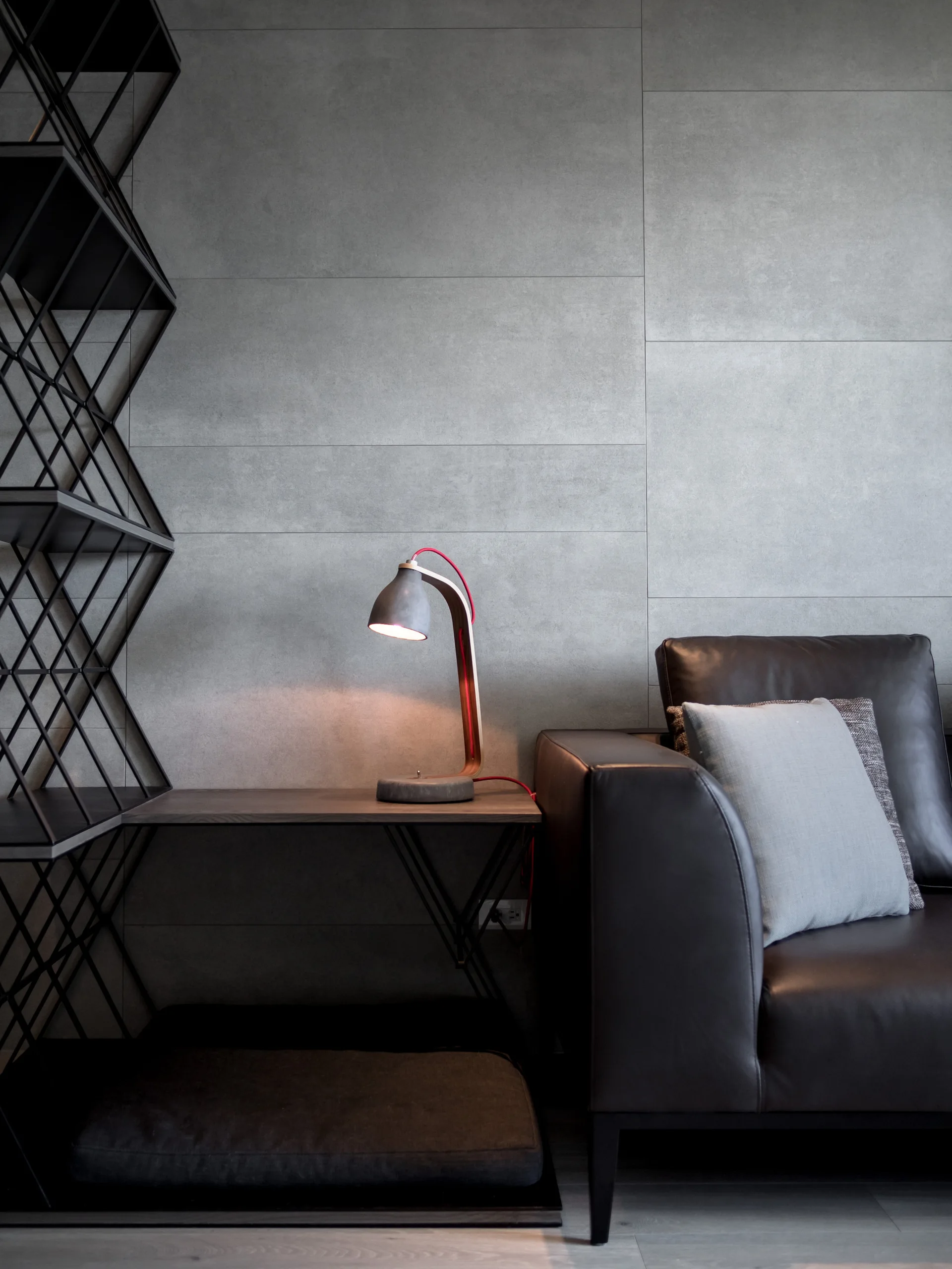 A modern living room features a dark leather sofa beside a wood side table with a concrete table lamp against a gray paneled wall.