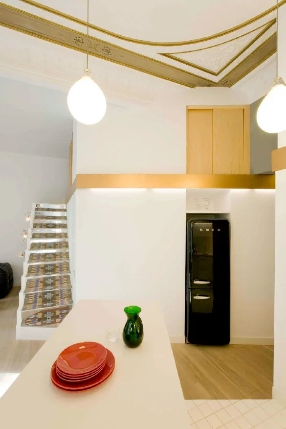 A modern kitchen with a white island, a recessed black refrigerator, an ornate gold-detailed ceiling, and a patterned tile staircase.