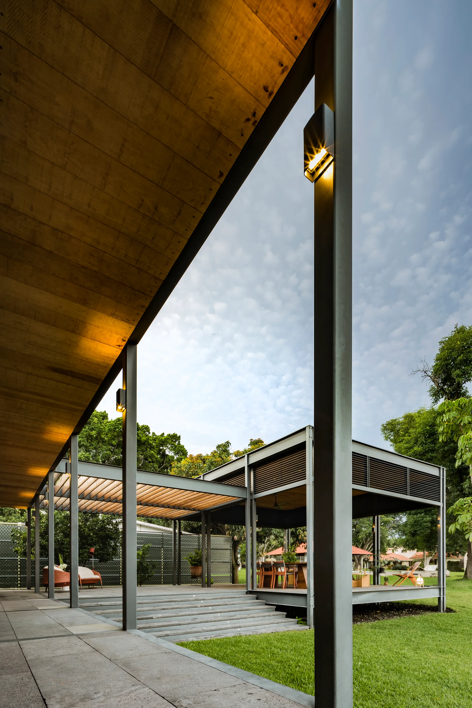 An open-air outdoor pavilion features a wooden plank ceiling, dark steel support beams, warm wall sconces, and stone steps leading to the lawn.
