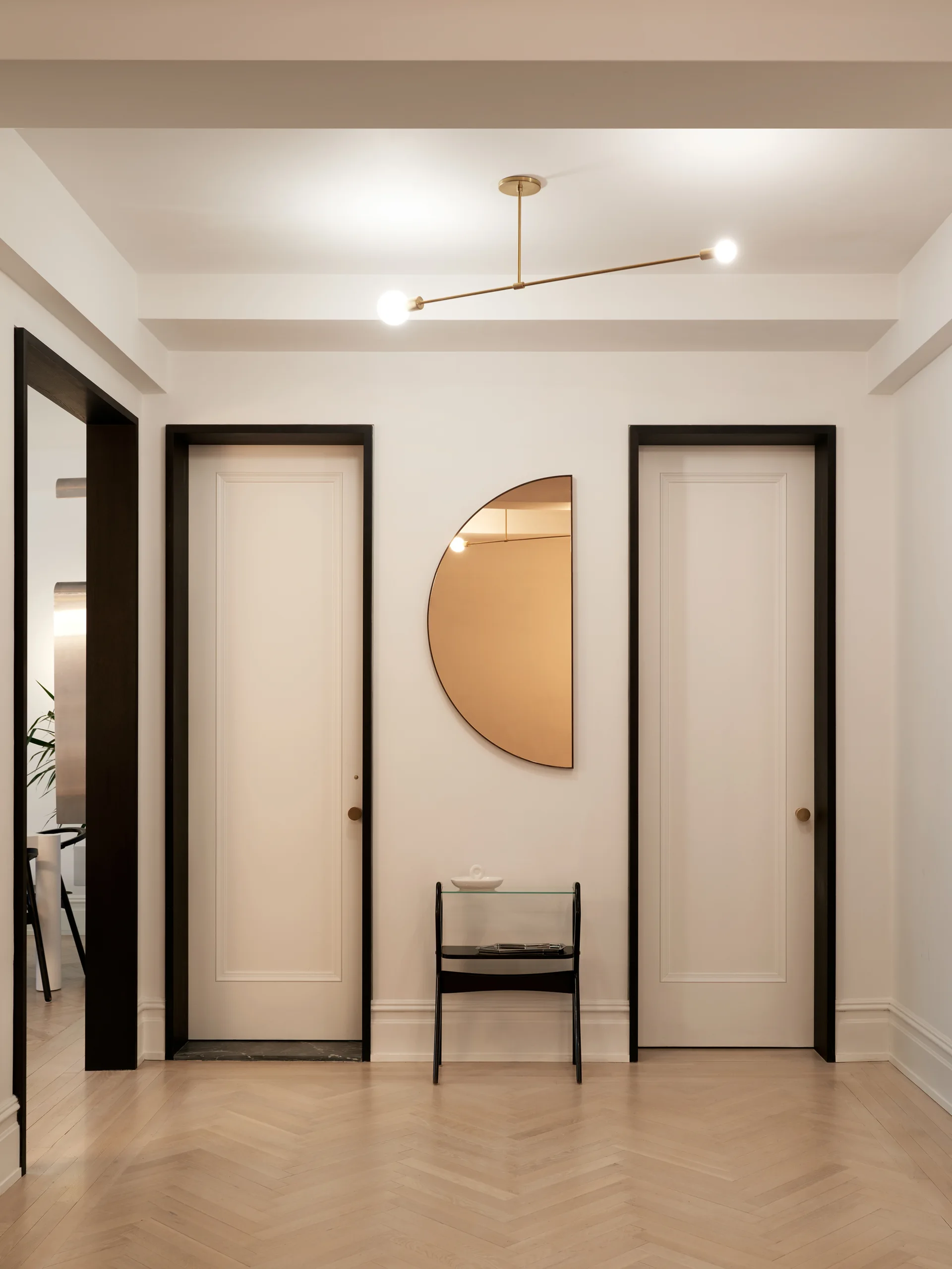 A minimalist residential hallway features two white doors with bold black trim, a light herringbone wood floor, and a semicircular mirror.