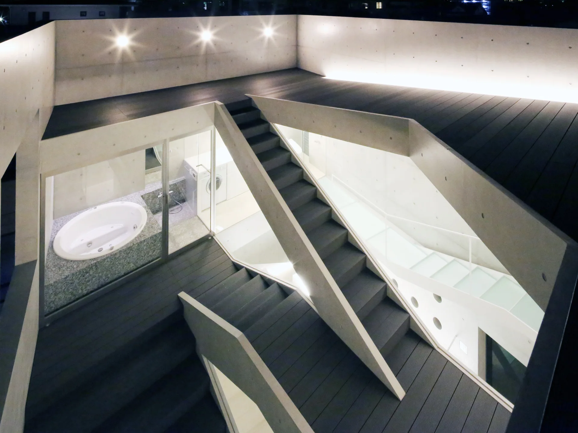 A multi-level outdoor concrete terrace features dark wood decking, a central geometric staircase, and glass walls showing an indoor bathroom with a tub.