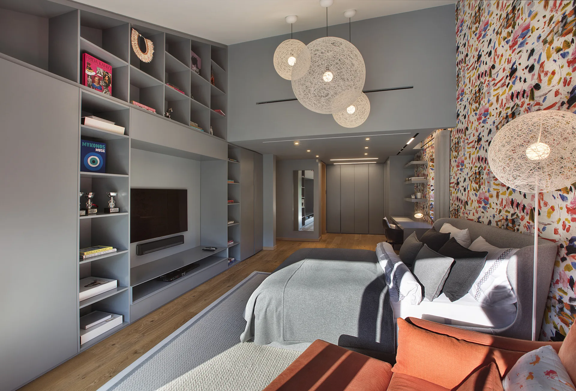 A contemporary bedroom features a grey upholstered bed, integrated grey shelving with a television, and vibrant multicolored abstract wallpaper.