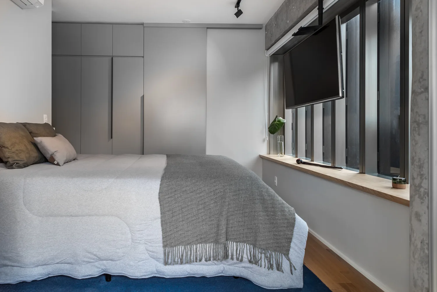 A minimalist bedroom with a grey made bed, flat-screen television, floor-to-ceiling grey closets, and a long wooden windowsill.