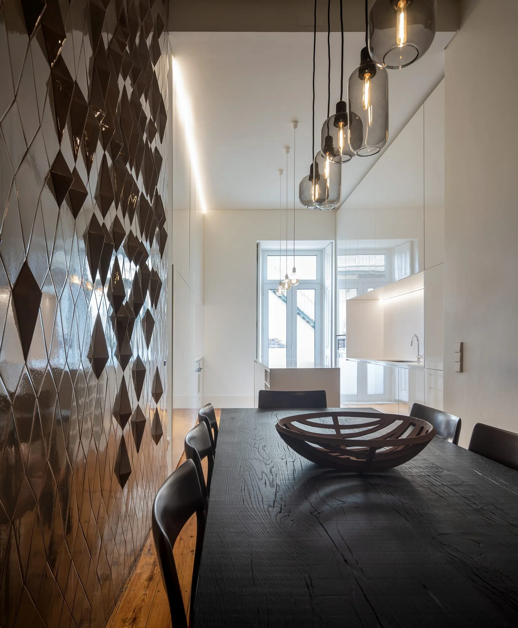 A modern dining room features a textured dark-brown geometric tile wall, a black wooden table, and hanging glass pendant lights.