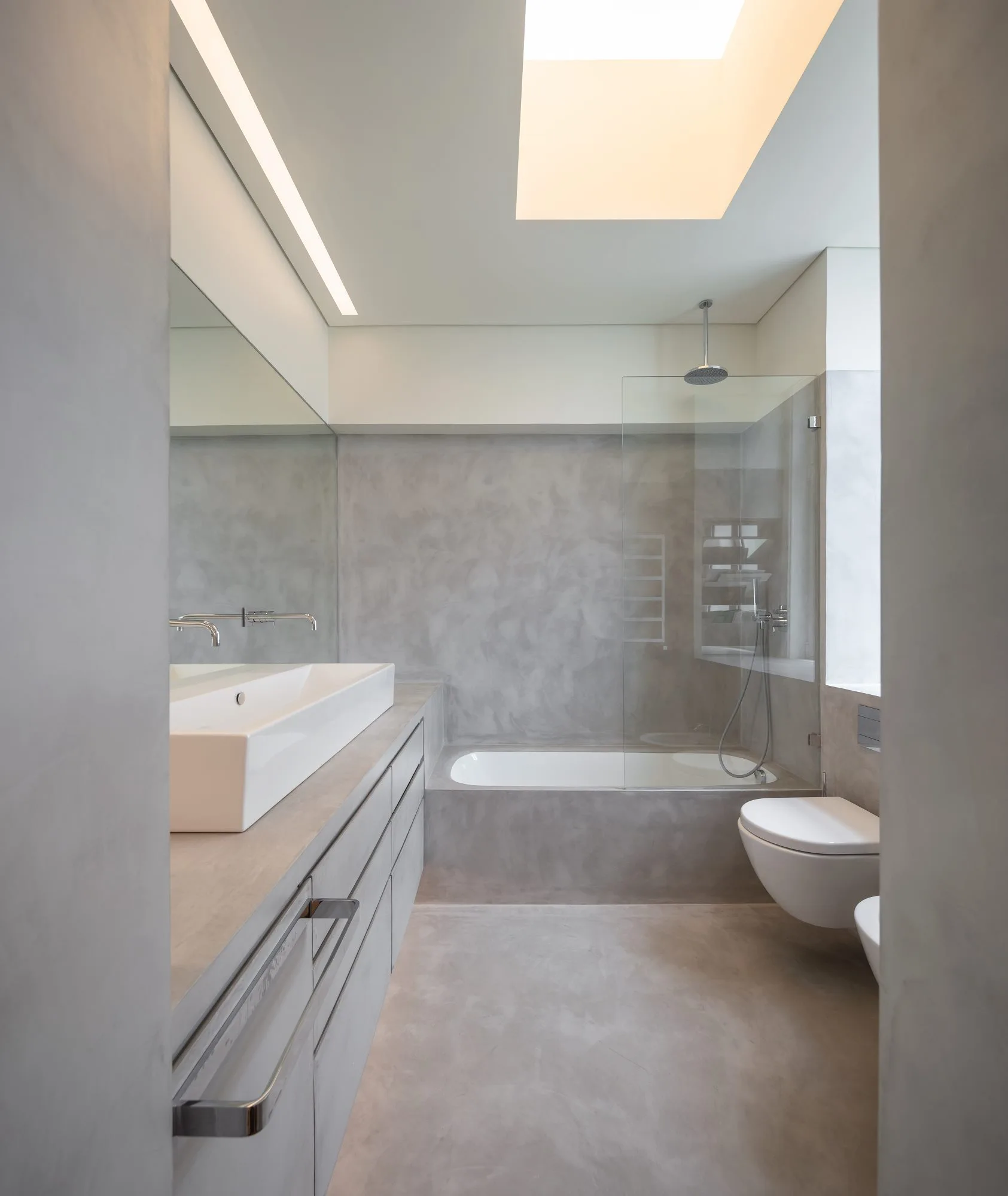 A minimalist bathroom featuring seamless grey concrete-like walls and floor, a white vanity sink, a wall-mounted toilet, and a large overhead skylight.