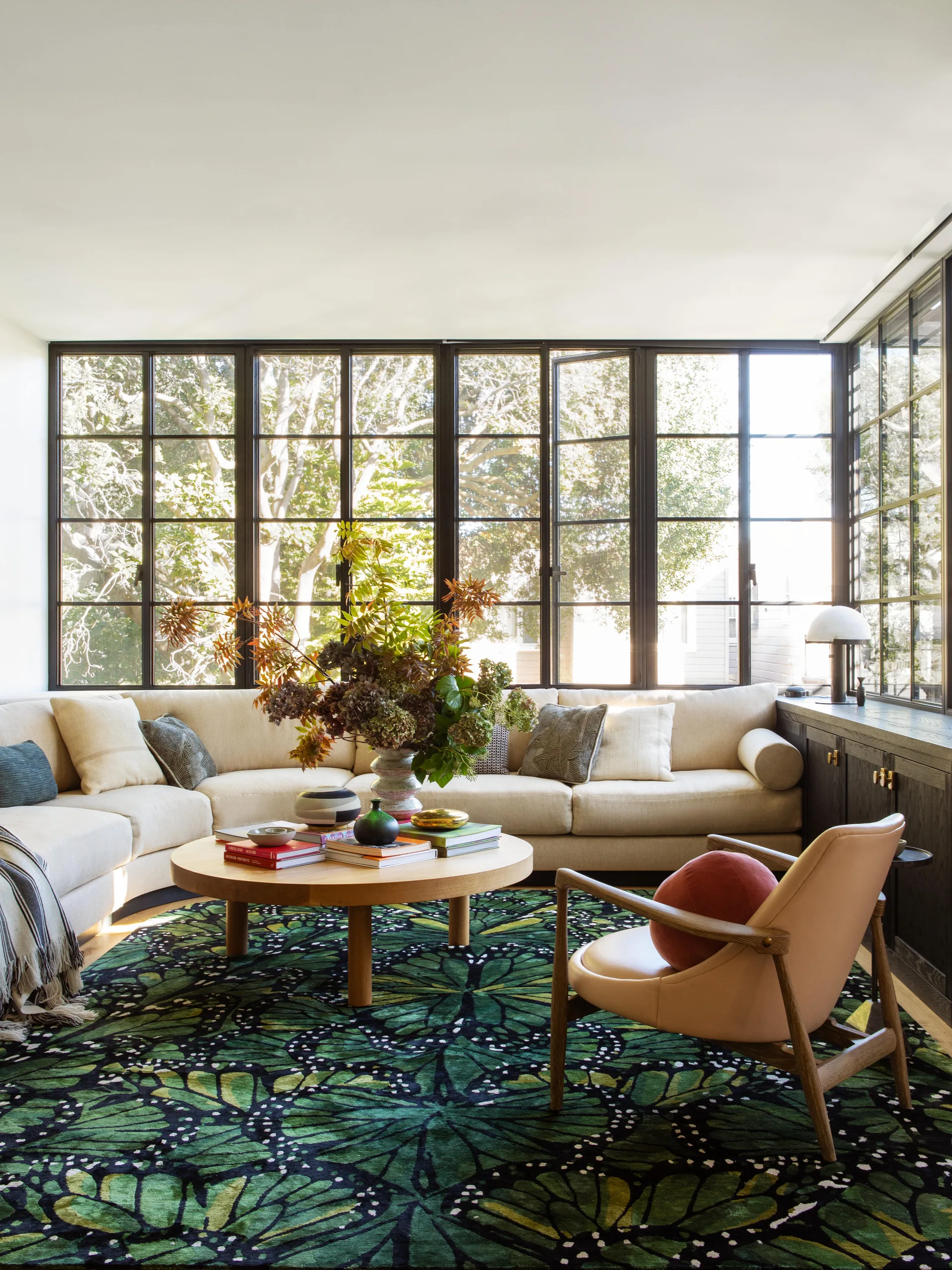 A light-filled living room features a cream sectional sofa, a round wooden coffee table, and a bold green patterned rug in front of large black grid windows.