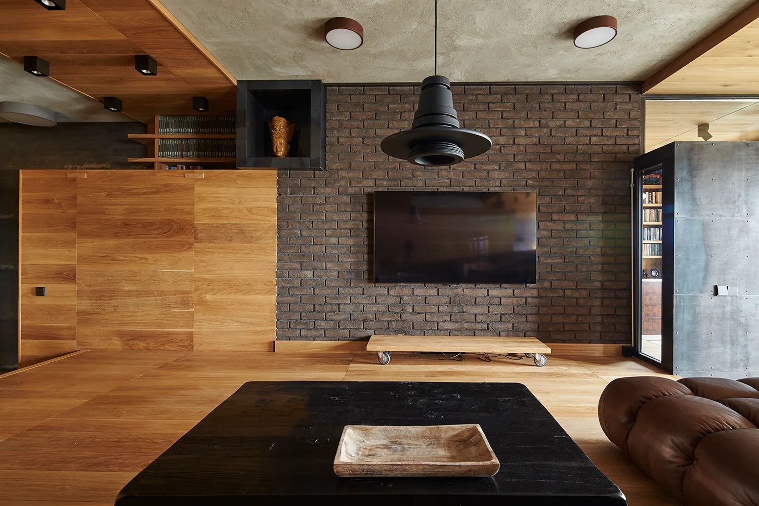 An industrial living room featuring a dark brick wall with a mounted TV, adjacent warm wood paneling, and a low wooden platform on casters.