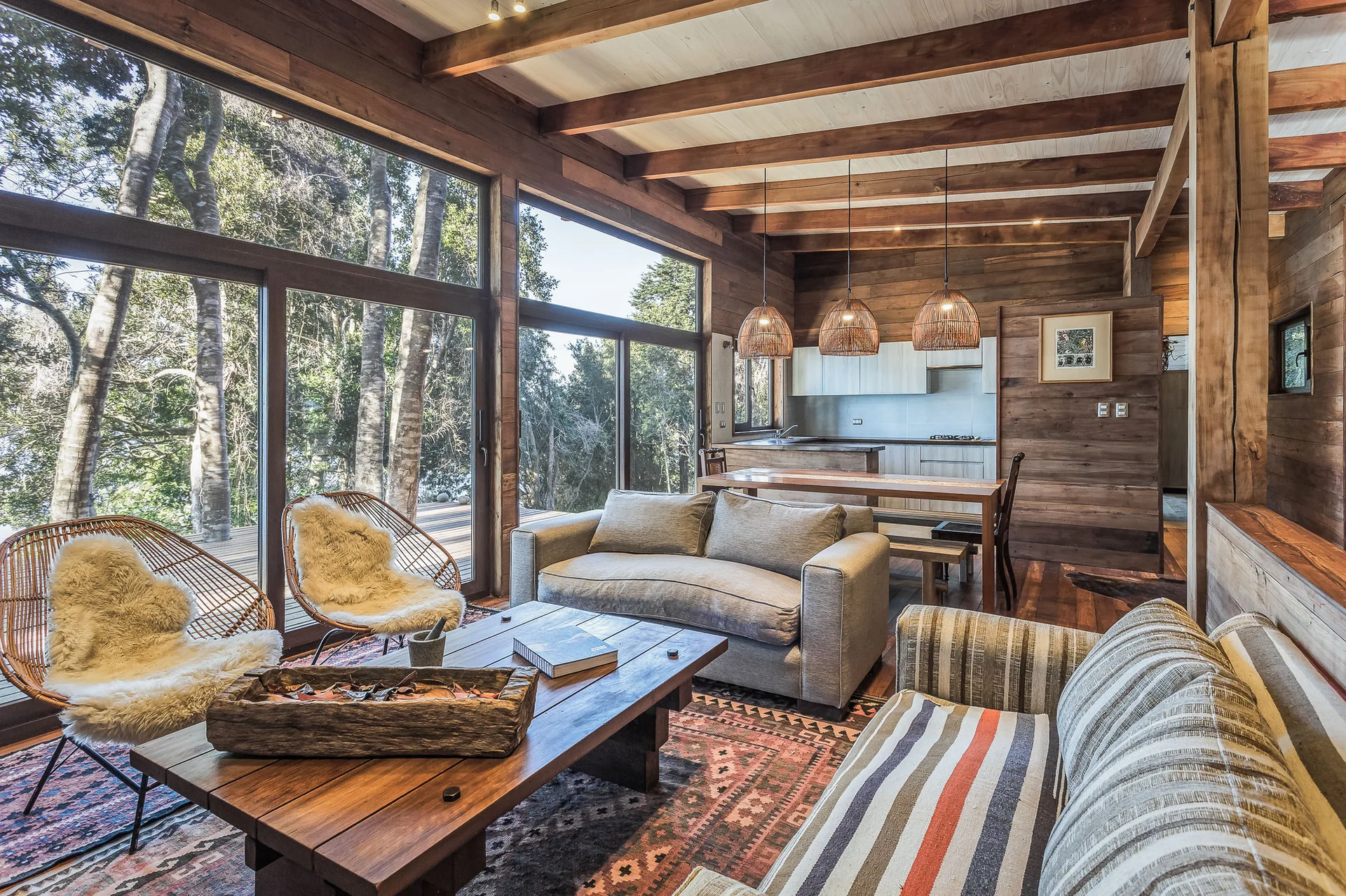 A rustic wood-paneled living room features comfortable sofas, woven accent chairs with faux-fur throws, and large windows looking out to a forest.