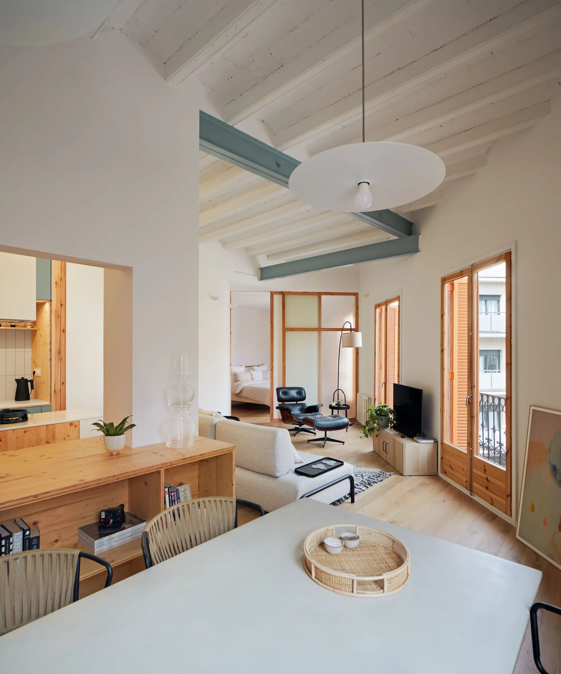 An open-plan living and dining room with white walls, light wood floors, exposed ceiling joists, and a teal accent beam.