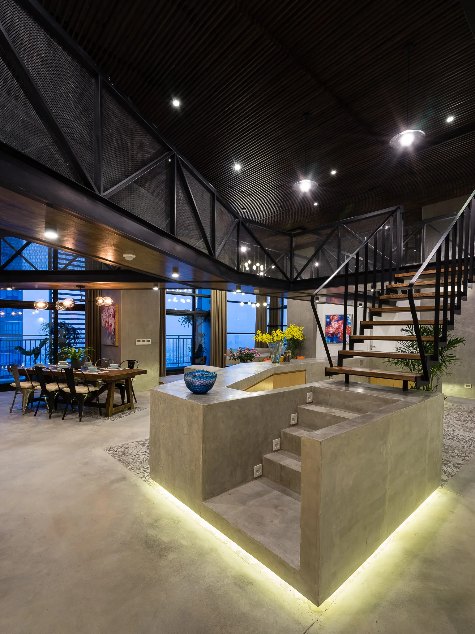 A central concrete staircase with wood steps and black railings leads to a black mesh mezzanine in an open-plan loft.