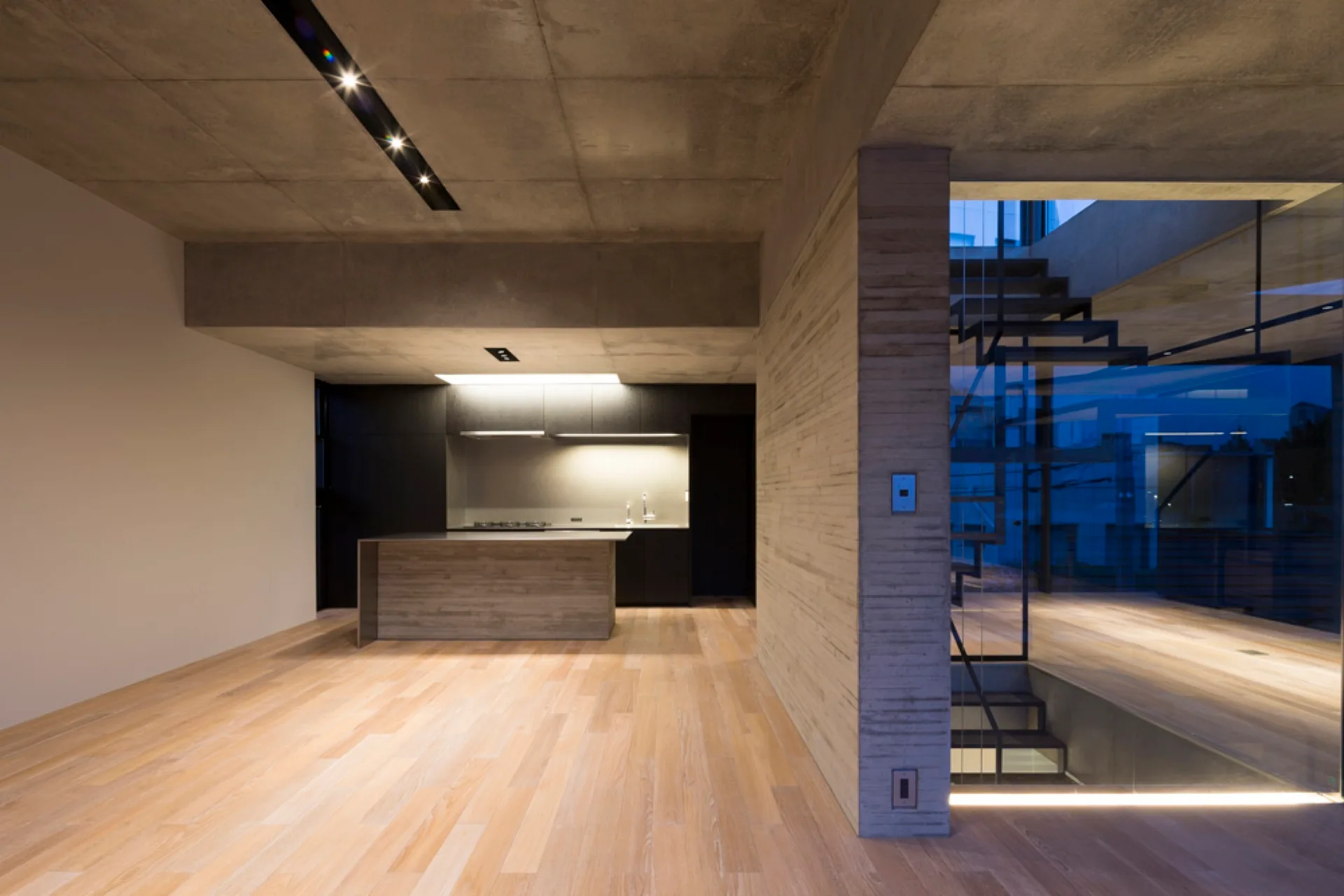 A modern minimalist kitchen with a board-formed concrete wall, light wood flooring, dark cabinetry, and a glass-enclosed staircase.