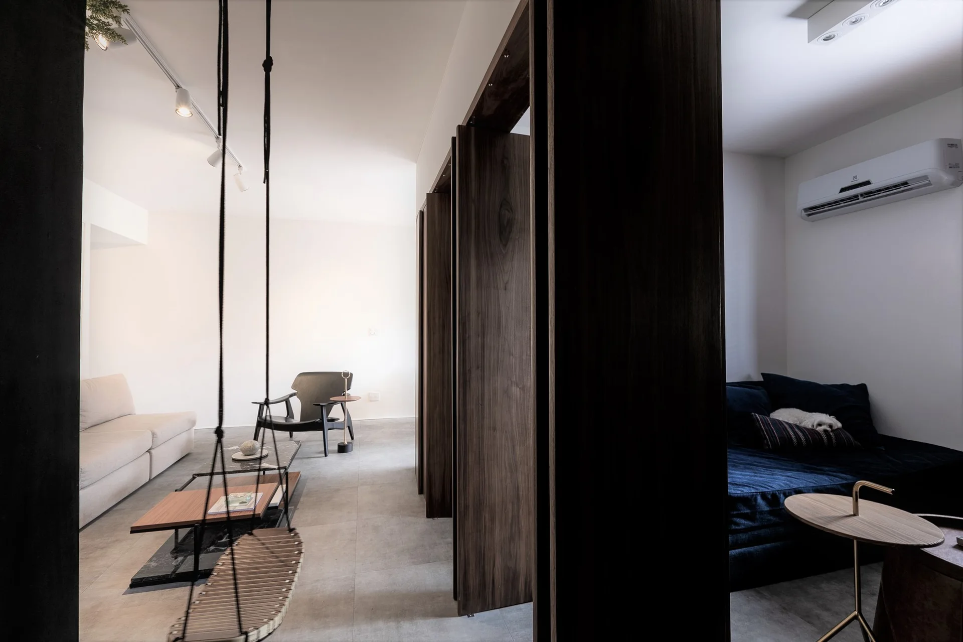 A minimalist apartment interior with a dark wood pivoting room divider separating a gray-tiled living area from a room with a navy daybed.