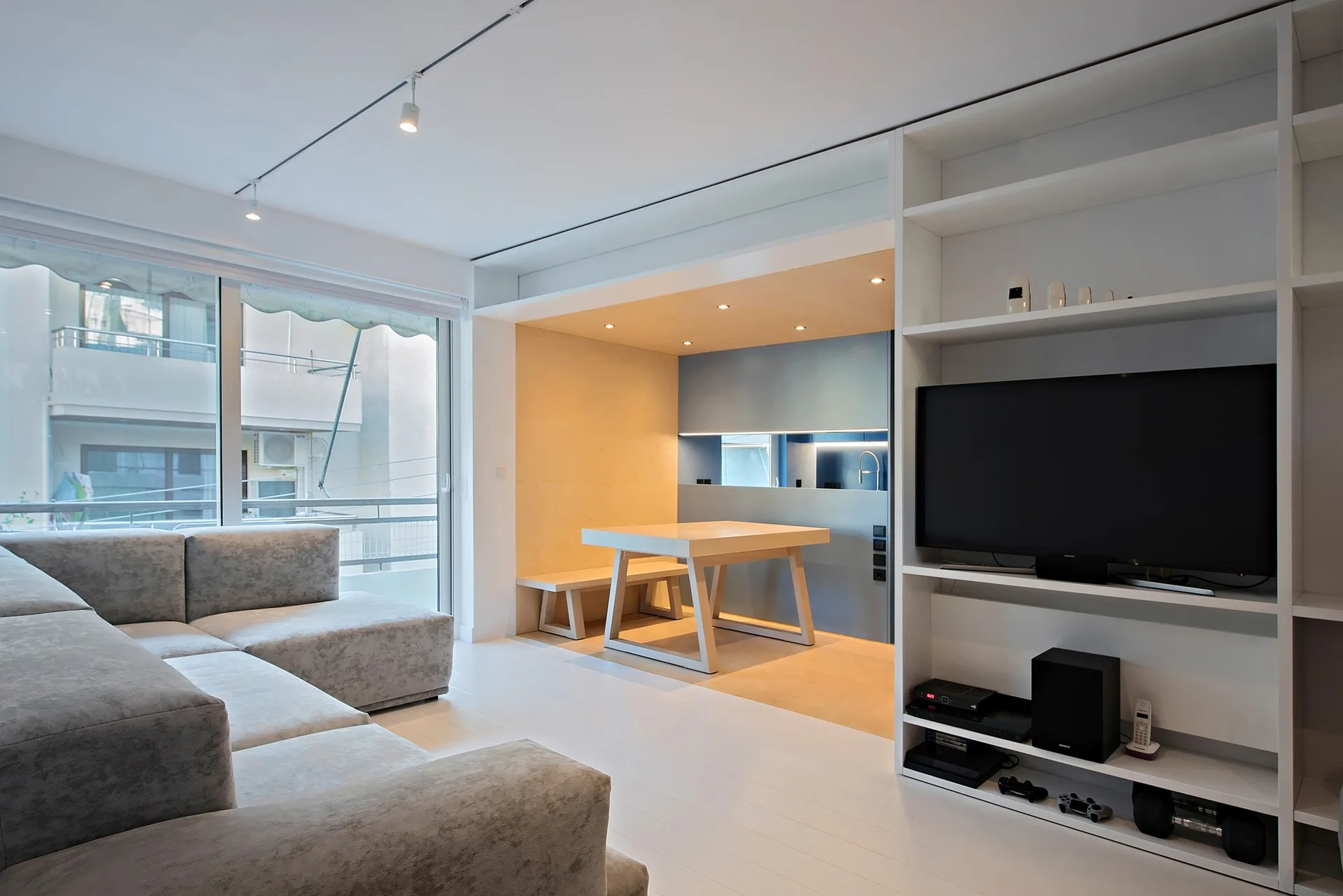 A white living room featuring a grey sectional sofa, a built-in media shelf, and a recessed wood-paneled dining alcove in the background.