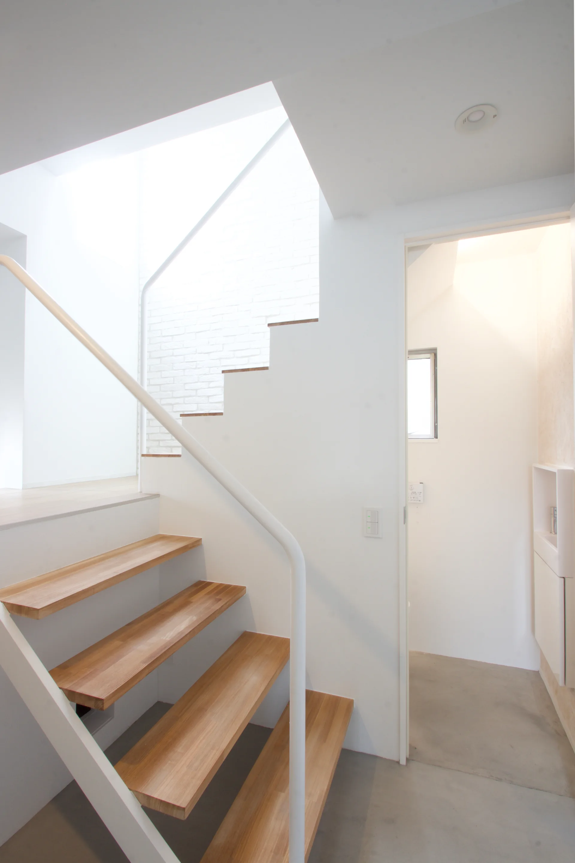 A minimalist staircase featuring open-riser wooden treads and a slim white metal handrail next to a concrete floor and white walls.