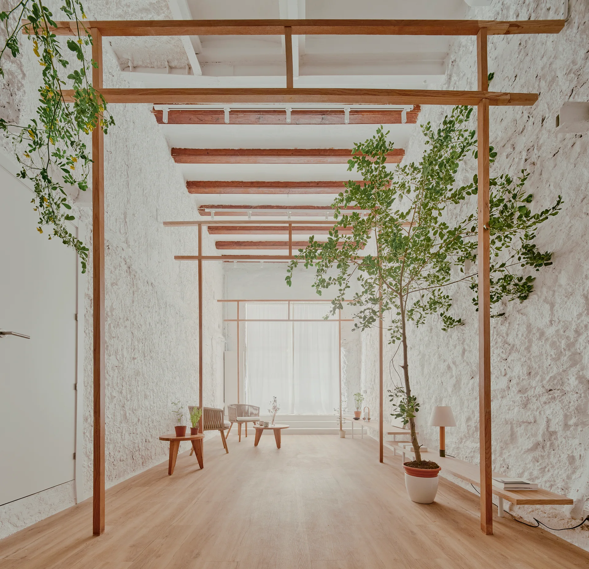 A minimalist room features heavily textured white plaster walls, light wood flooring, exposed ceiling beams, and slim wooden frames acting as dividers.