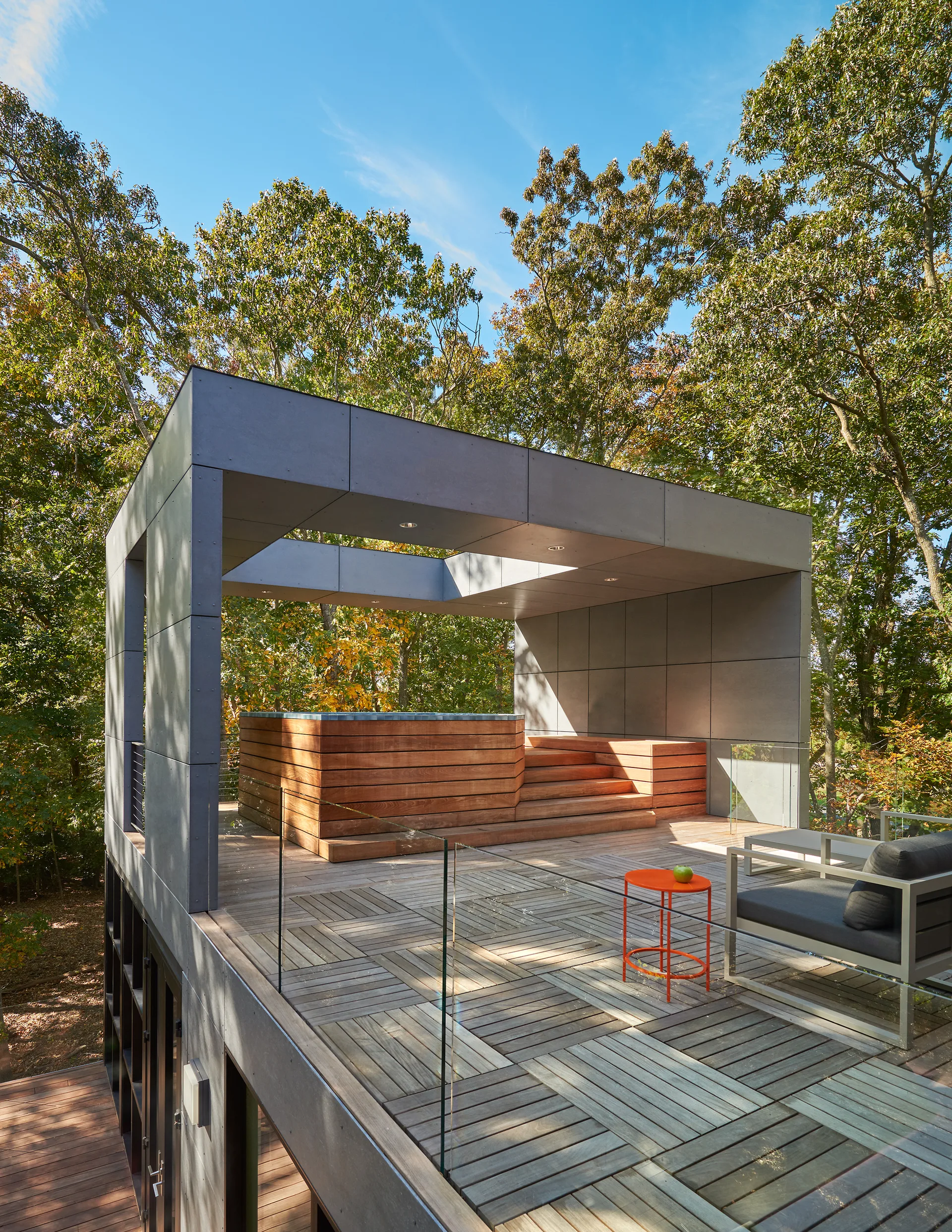 A modern outdoor wooden deck features a wood-clad hot tub under a grey architectural canopy alongside a glass railing and minimalist seating.