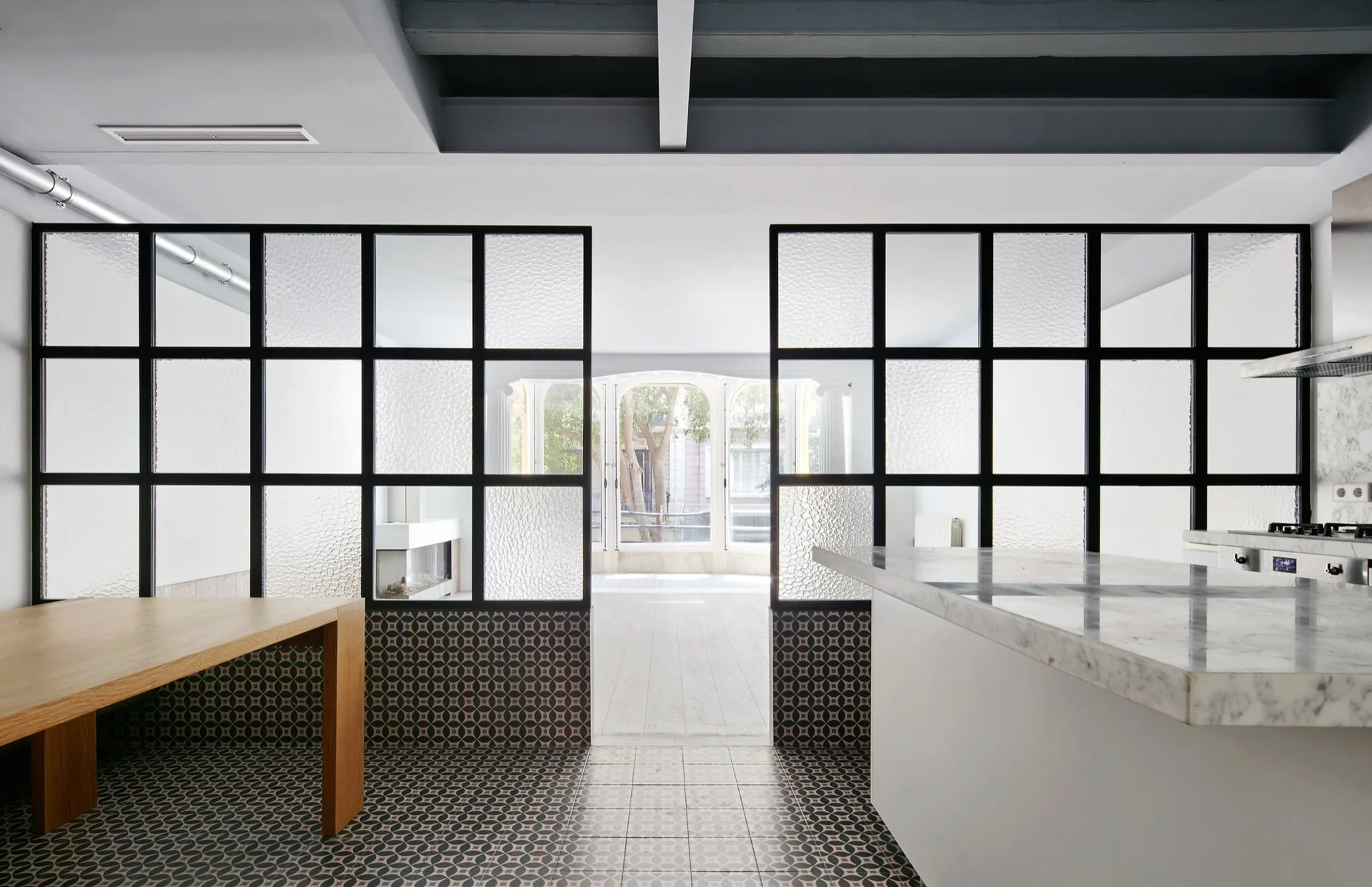 A modern kitchen featuring a marble island and wooden table, separated from an adjacent room by a black-framed textured glass partition over patterned tiles.