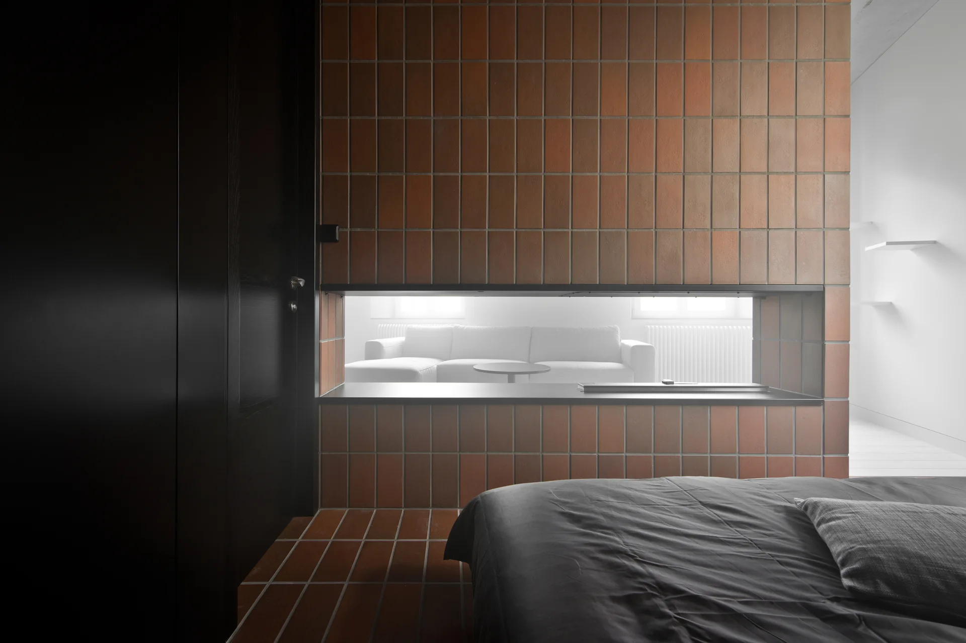 A view from a bedroom looking through a horizontal slot in a terracotta tiled wall partition toward a bright living room with a white sofa.