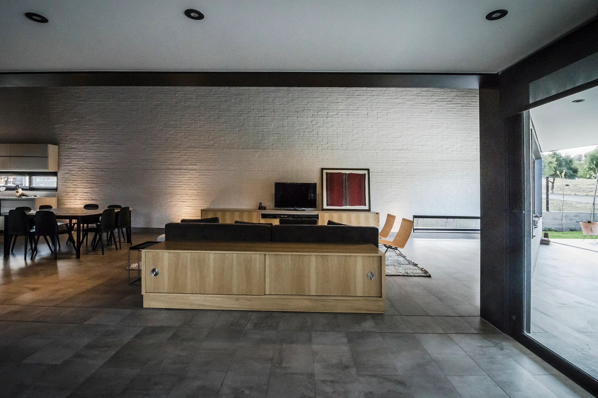 An open-plan living room features a painted white brick wall, a low wooden sofa console, a dark dining set, and grey tile flooring.