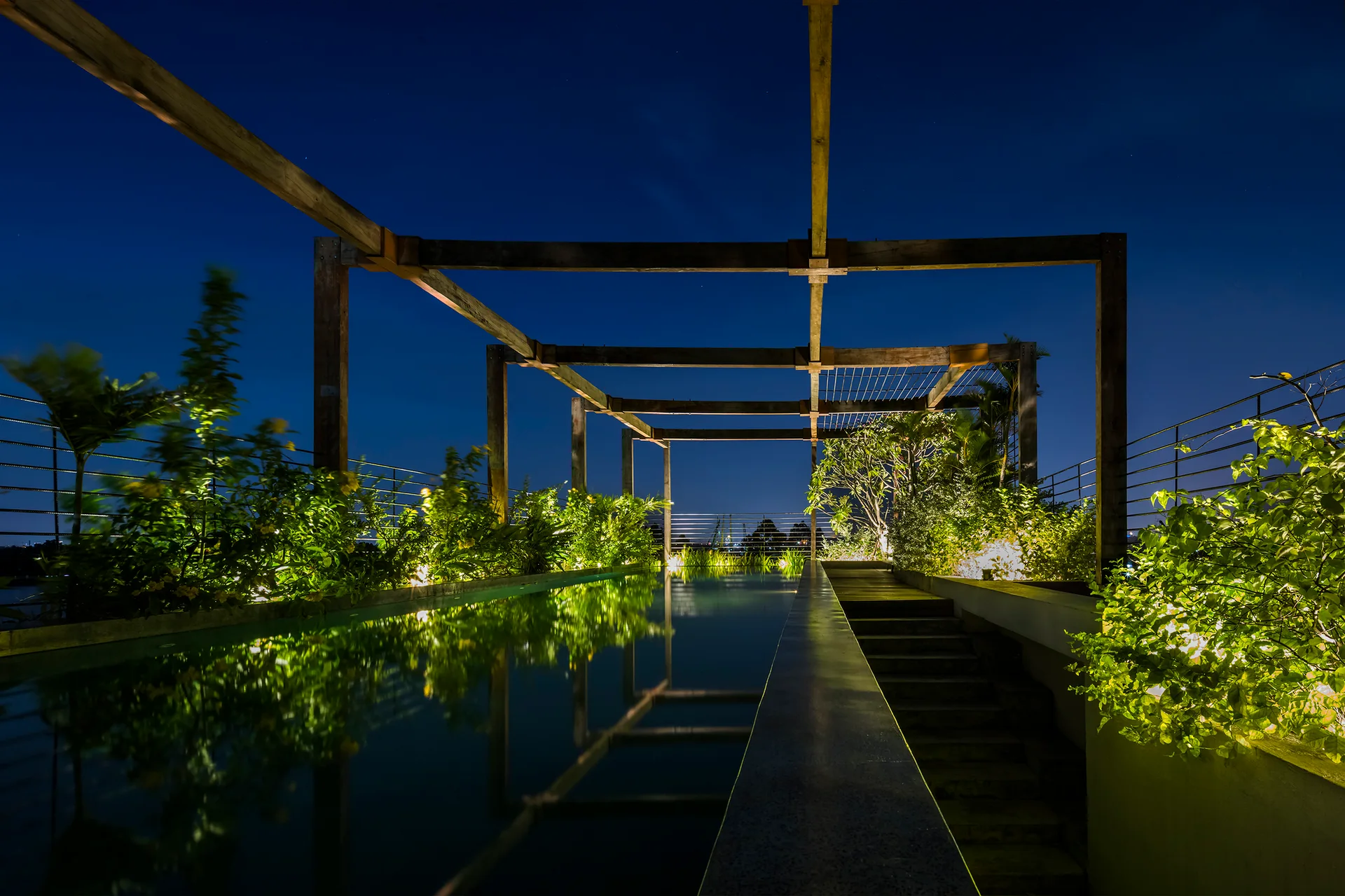 An elongated outdoor lap pool at night framed by a tall wooden timber pergola, flanked by illuminated planters and stone steps.