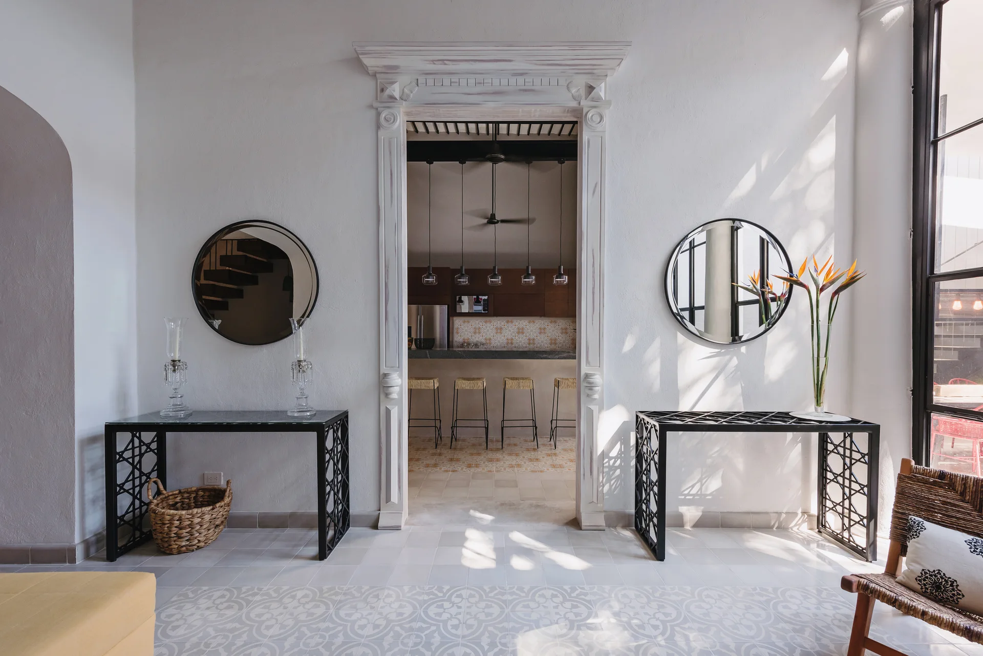 Symmetrical black metal console tables and round mirrors flank an ornate white doorway leading into a kitchen with patterned tiles.