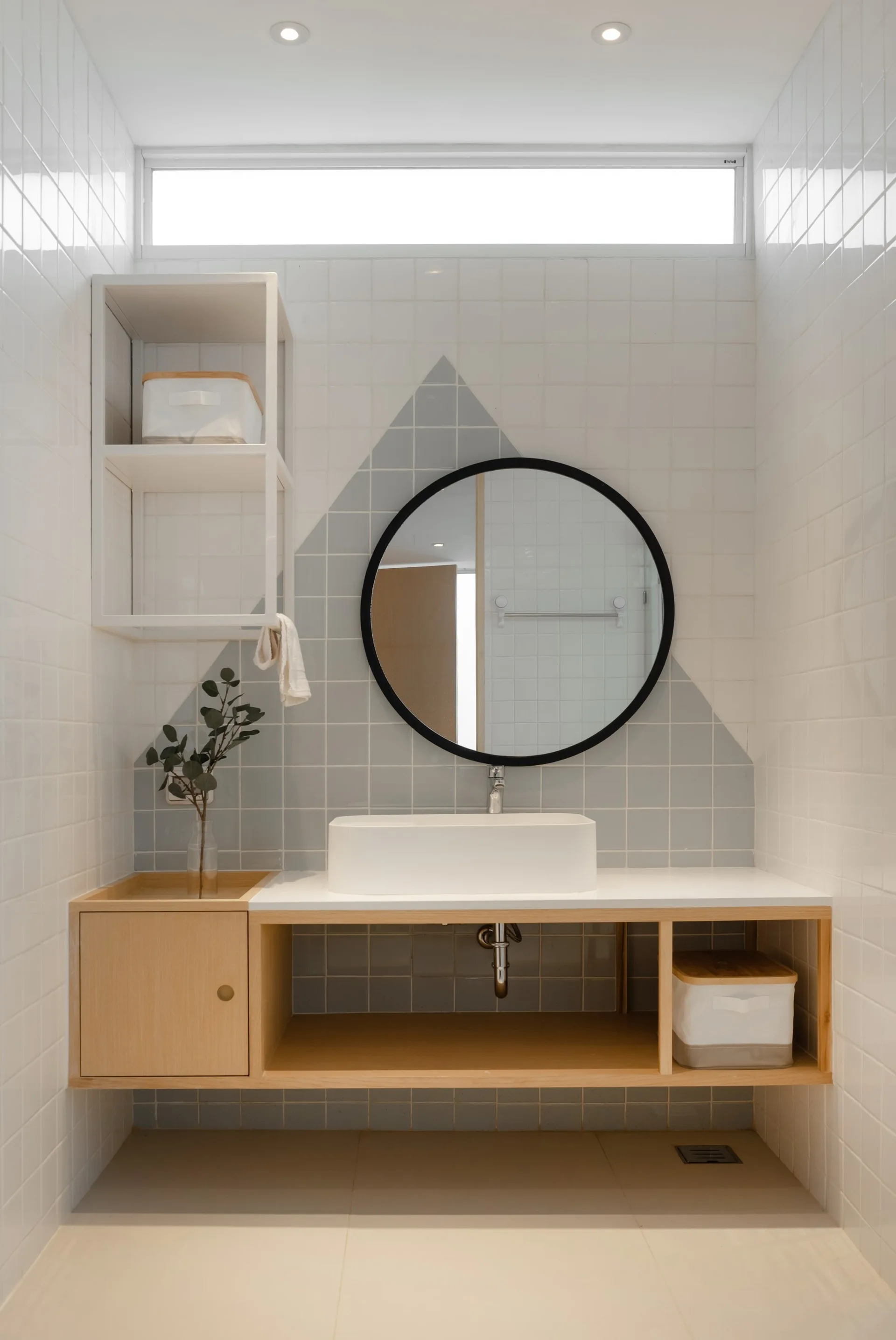 A light wood bathroom vanity with a vessel sink is set against a white tiled wall featuring a grey geometric triangle pattern and a round mirror.