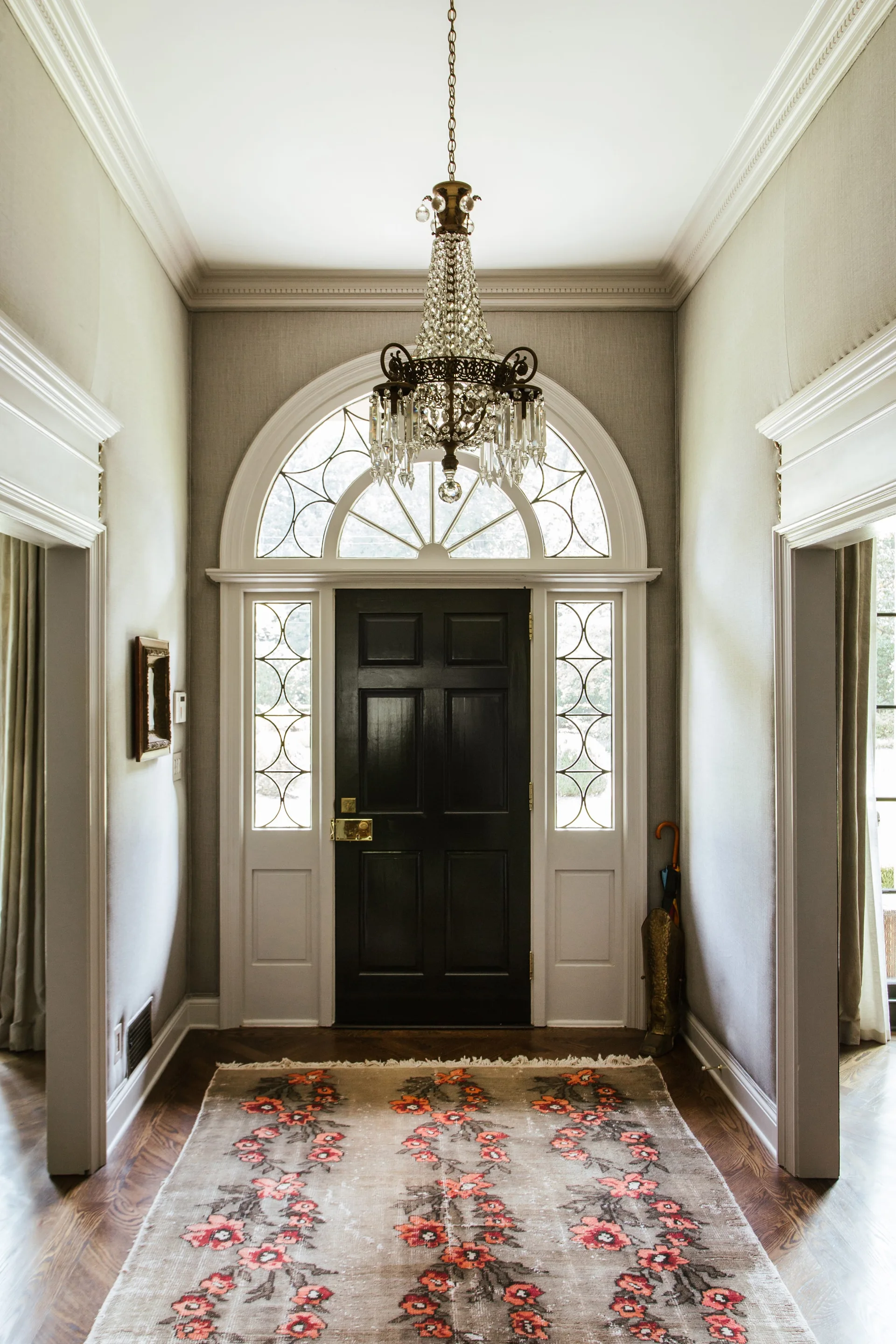 A residential foyer featuring a black paneled front door with an arched fanlight, sidelights, a crystal chandelier, and a patterned runner rug.