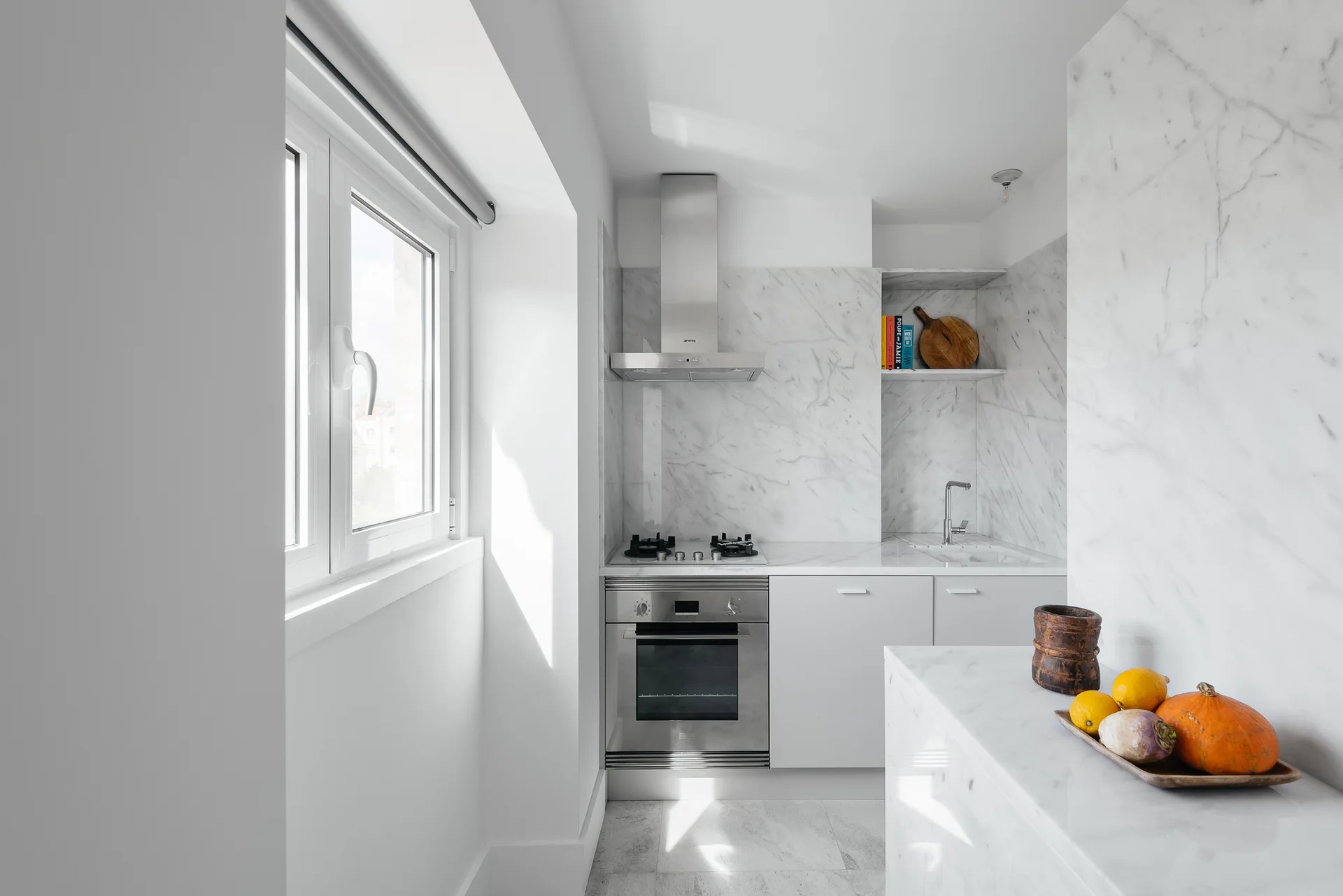 A minimalist galley kitchen with white flat-front cabinets, a stainless steel stove, and white marble slab countertops and backsplash.
