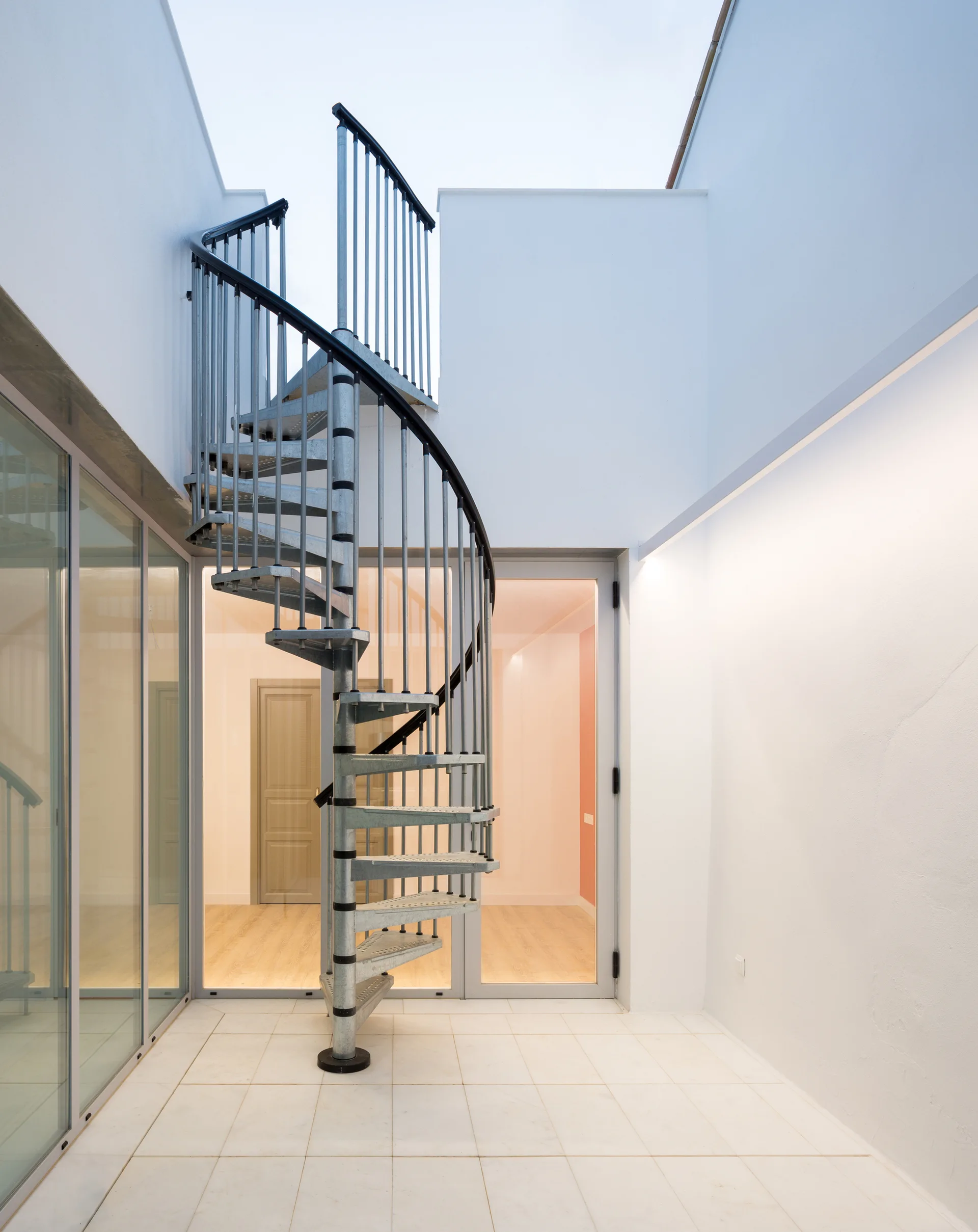 A galvanized steel spiral staircase stands in a minimalist white-walled courtyard with glass walls and light-tiled flooring.