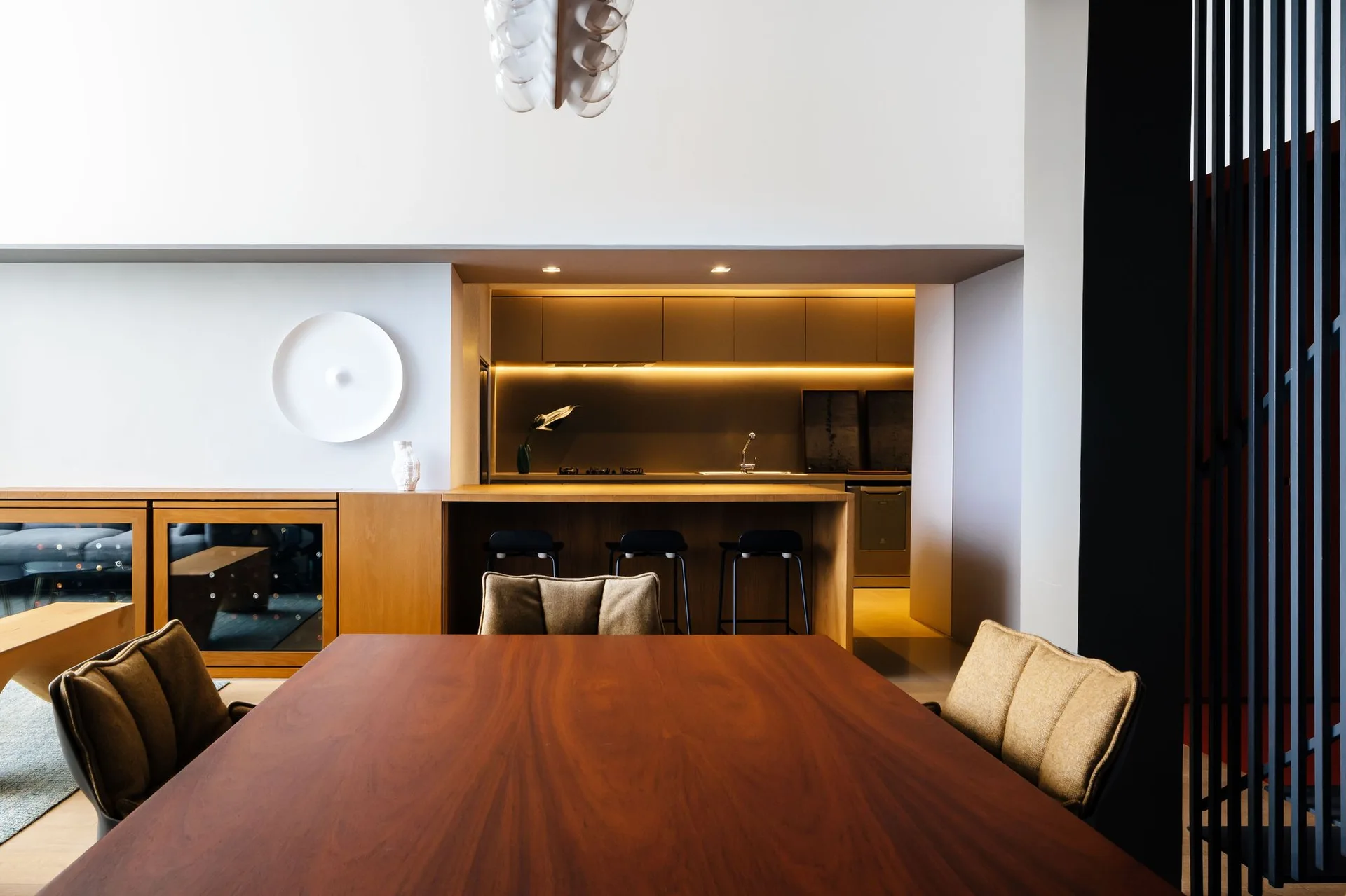 A spacious dining area features a large warm wood table in the foreground, leading to a recessed kitchen alcove with integrated cabinetry and bar stools.
