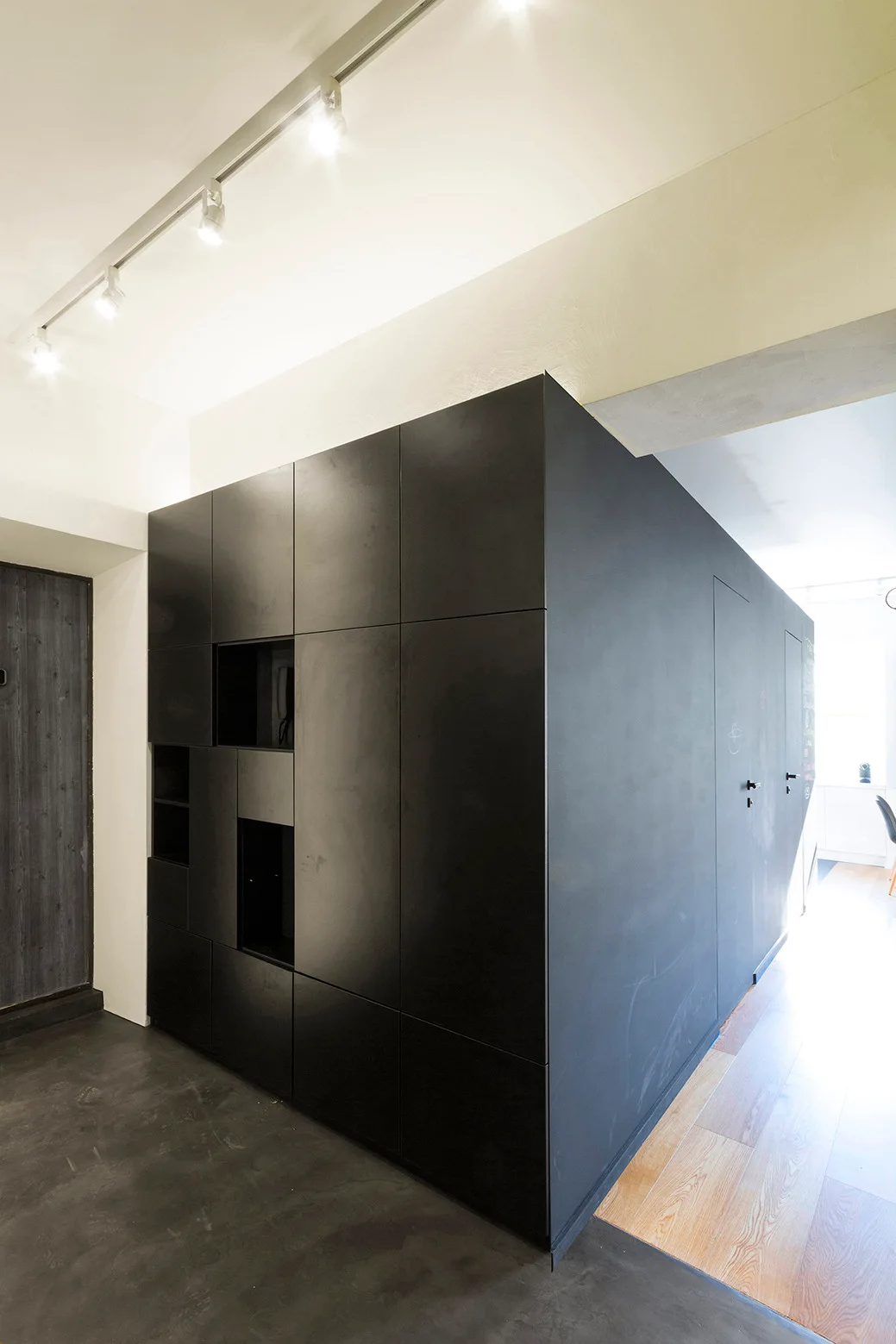 A large matte black architectural storage block with recessed niches and flush doors stands between a dark floor and wood flooring.