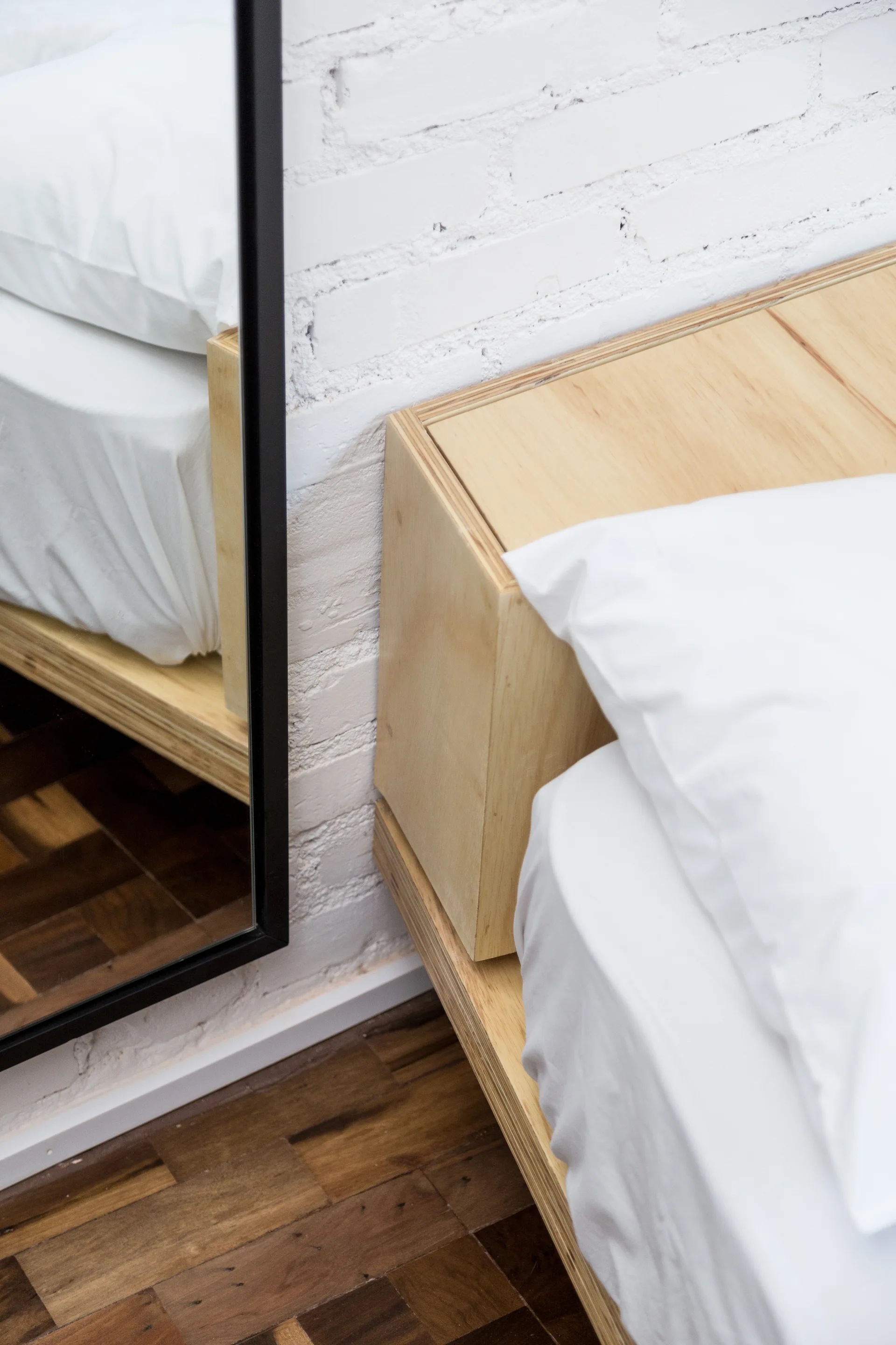 A close-up of a light plywood bed frame and white pillow next to a tall black-framed mirror against a white painted brick wall.