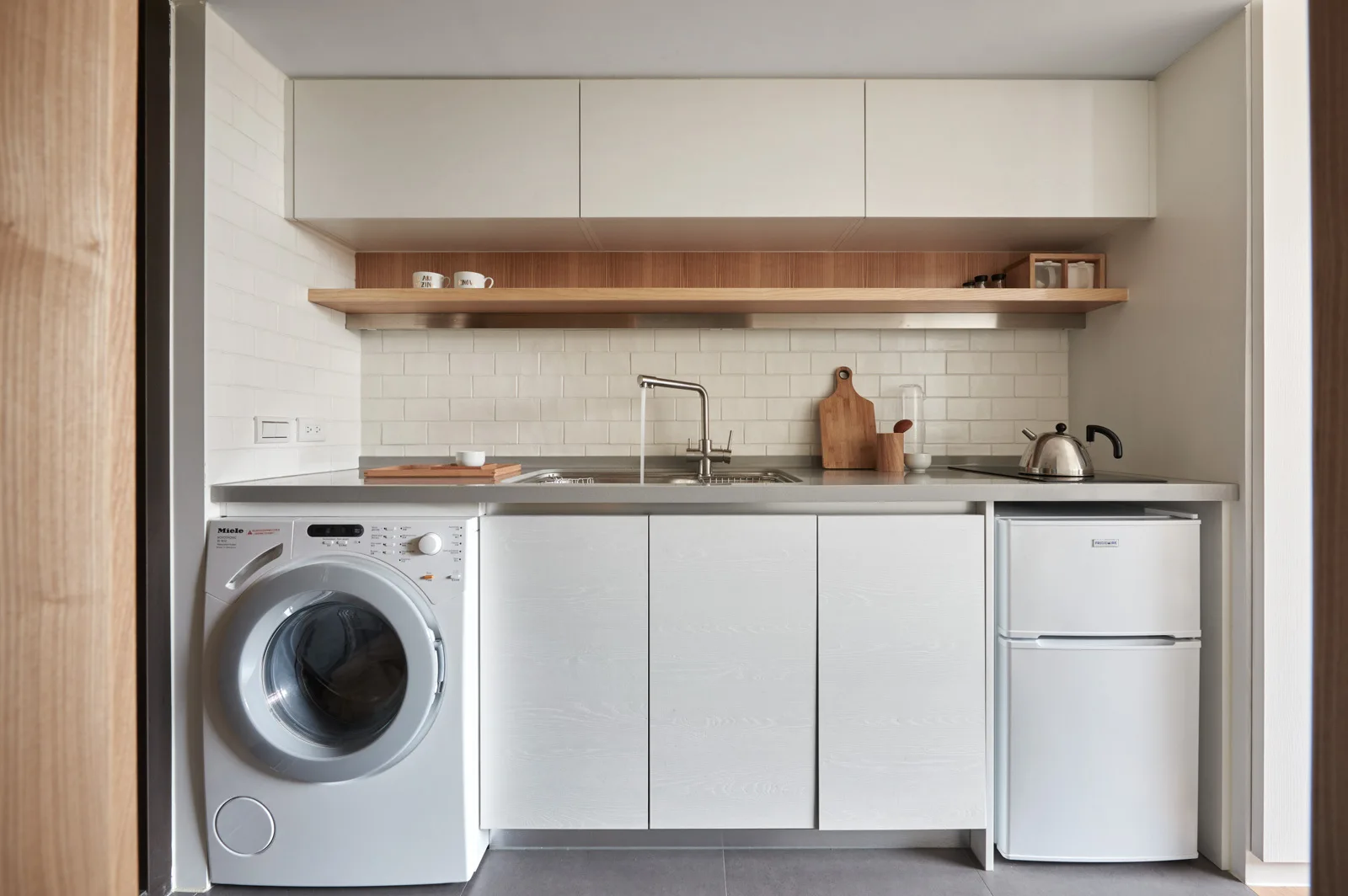 A minimalist white kitchenette featuring integrated appliances, a small sink, a light wood floating shelf, and a white subway tile backsplash.