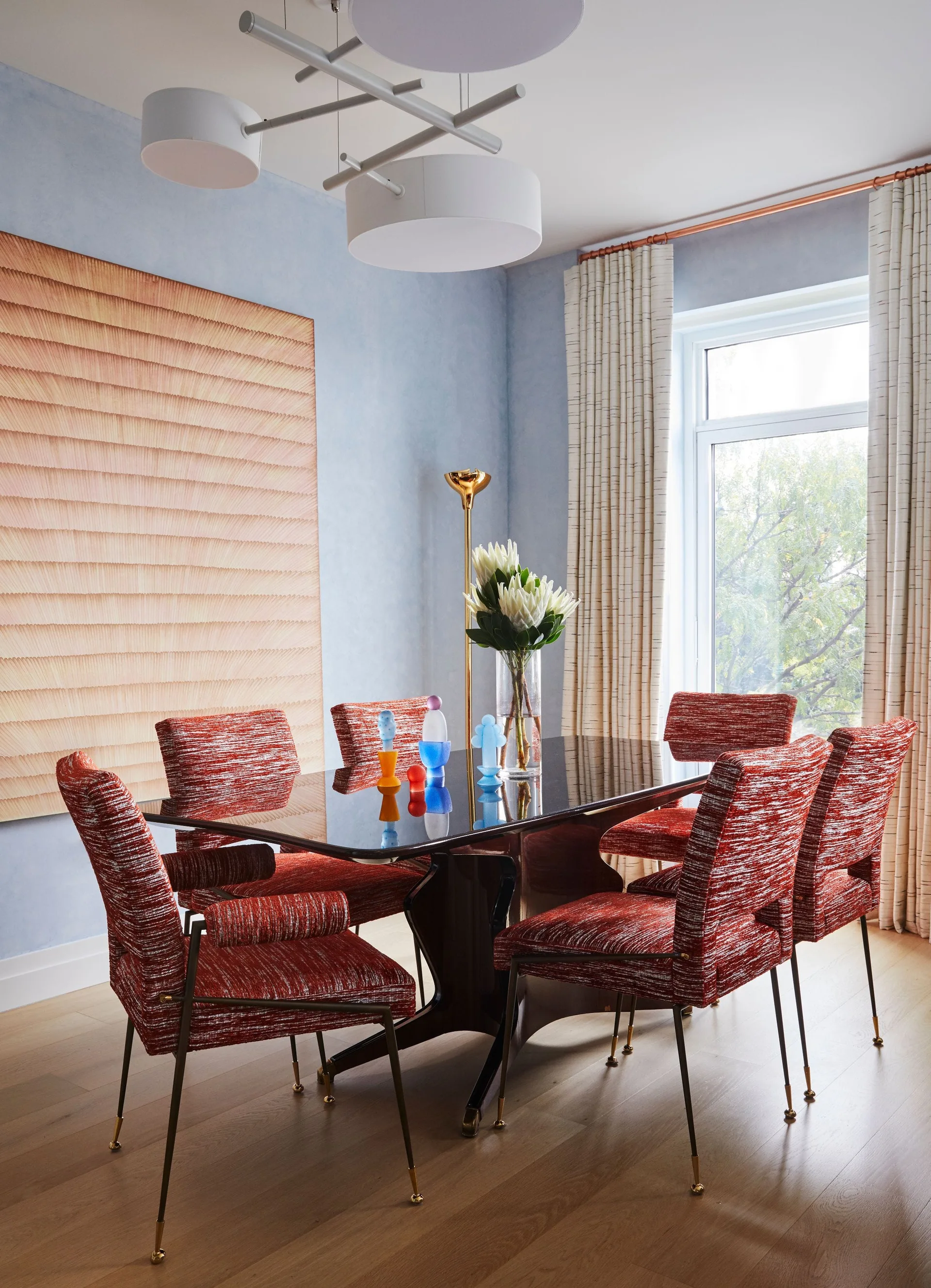 A modern dining room features a glossy black table, textured red upholstered chairs, and light blue walls with a large abstract canvas.