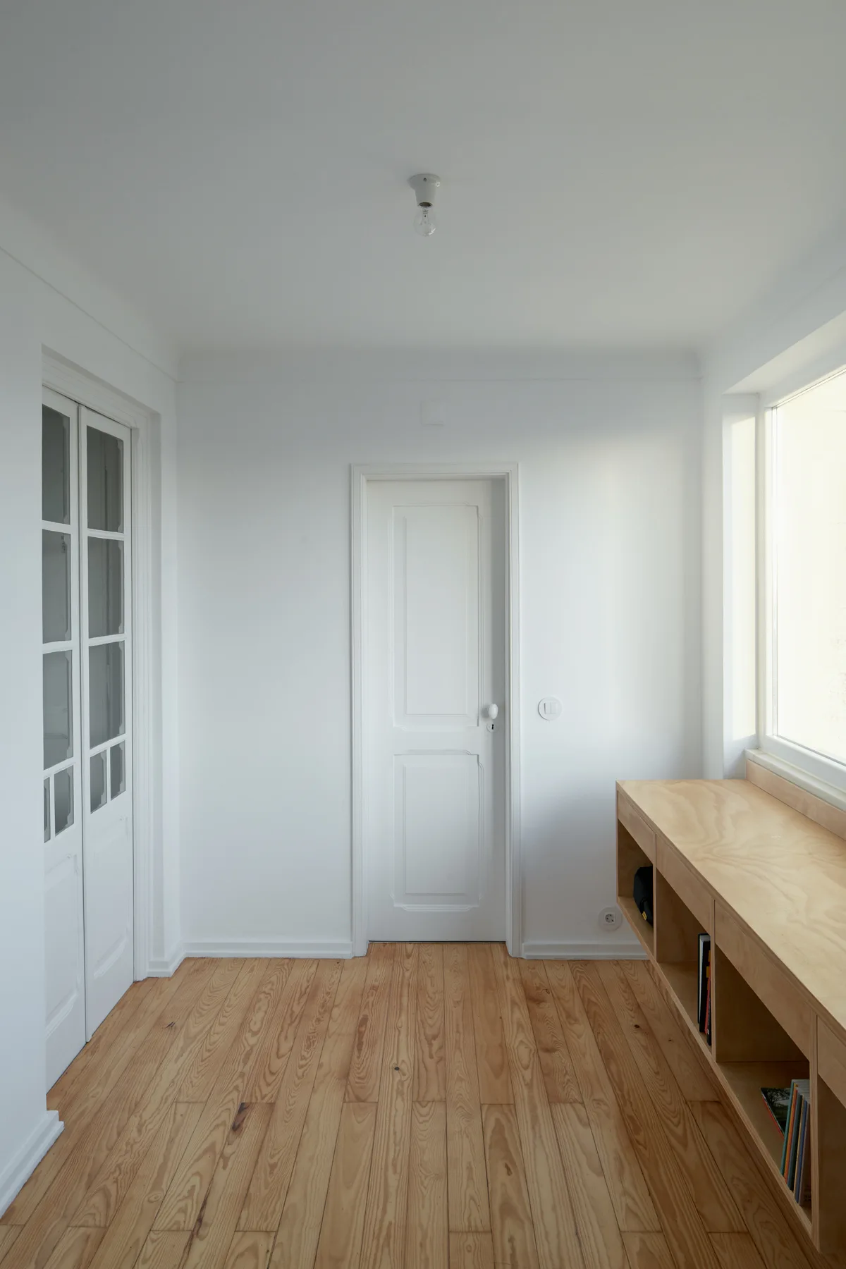 A minimalist white hallway features light wood floors, glass-paned French doors, and a custom low-profile plywood shelving unit under a window.